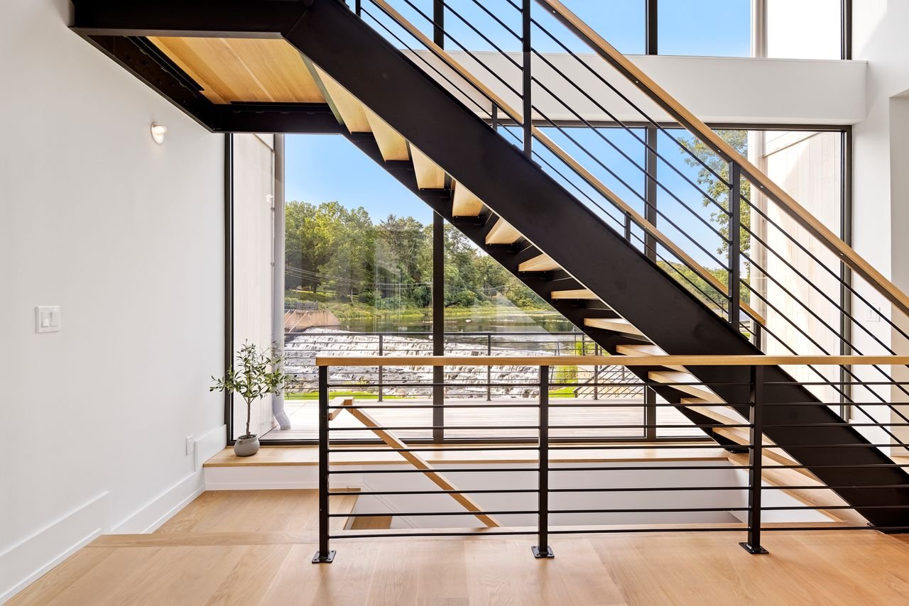Interior view of a modern house with a staircase featuring black metal framing, wooden steps, and a sleek wooden handrail, leading to an upper level with large floor-to-ceiling windows celebrating natural light and outdoor scenery.