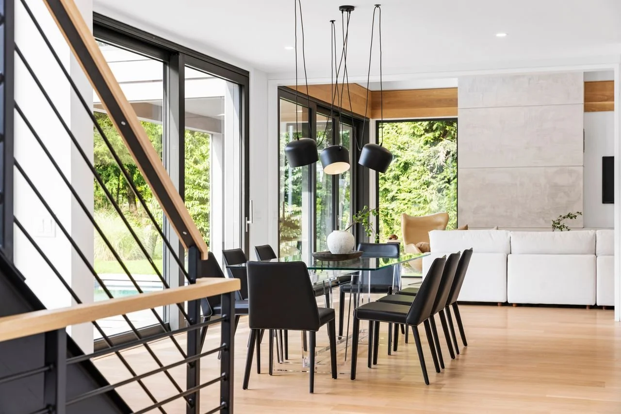 Modern living room with a glass dining table, black chairs, and large windows showing greenery outside