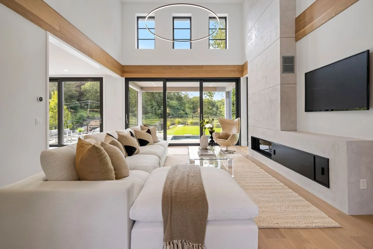 Modern living room with white sofa, beige cushions, glass coffee table, beige armchair, large sliding glass doors opening to a green yard, high ceiling with circular chandelier, and a mounted flat-screen TV on a white stone wall.