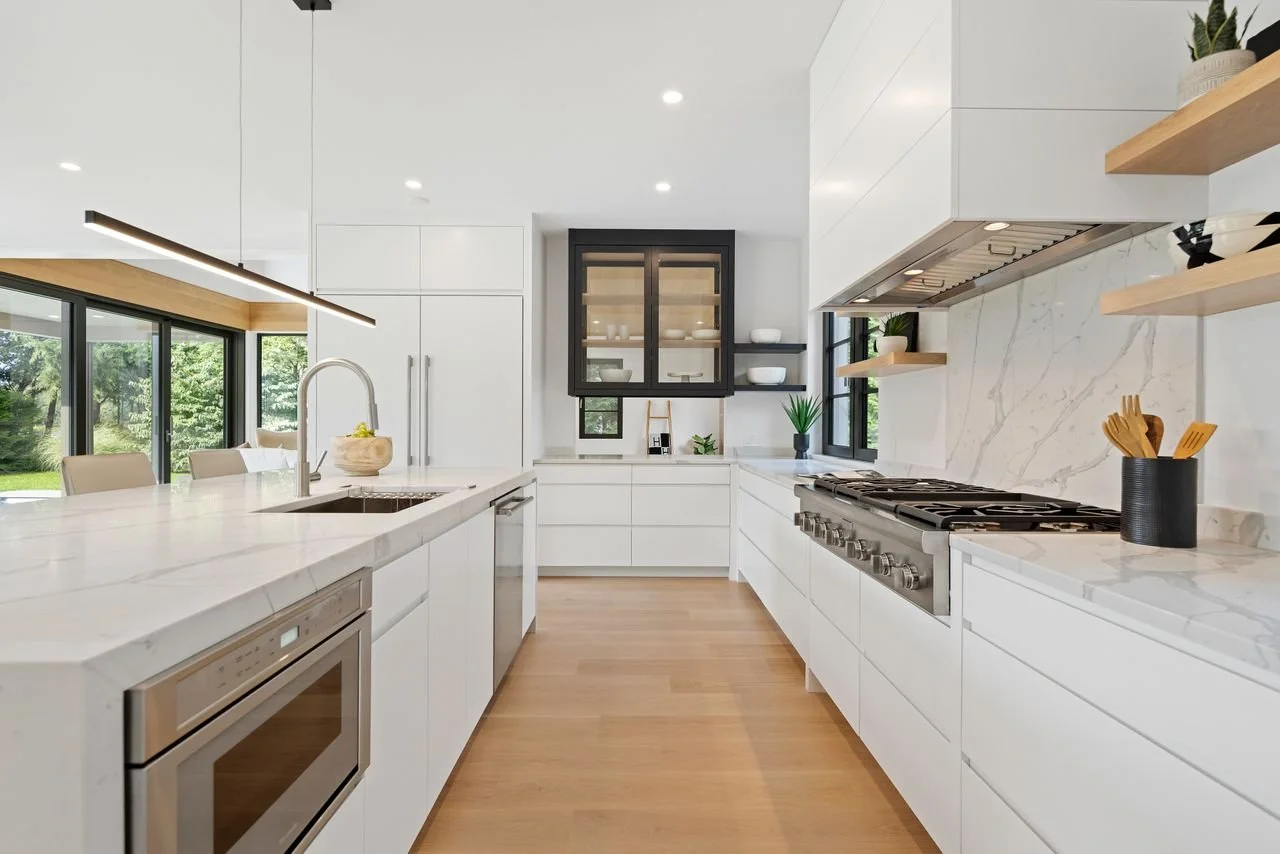 Modern kitchen with white cabinets, marble countertops, black window frames, and wooden shelves. Features a gas stove, microwave, and a view of green trees through sliding glass doors.