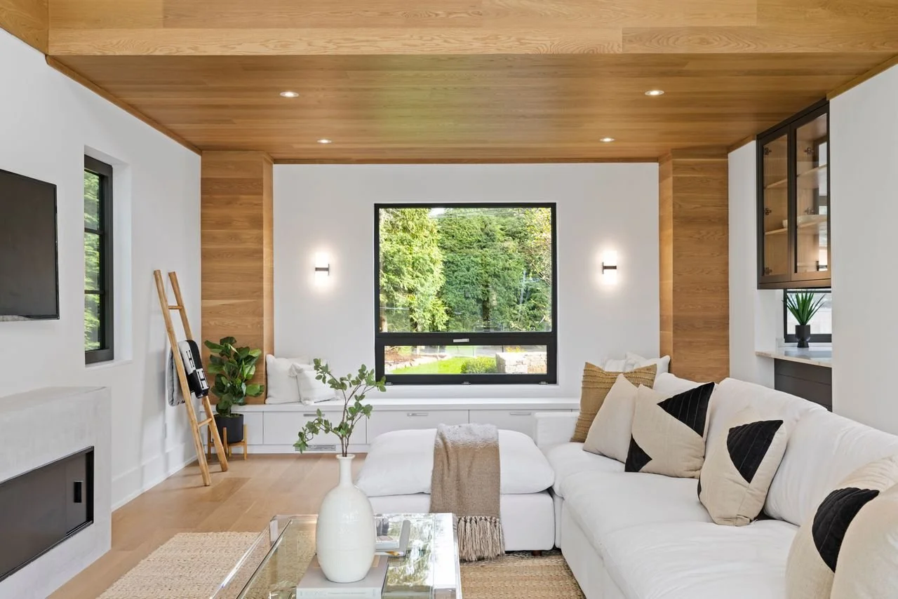 Modern living room with white walls, wooden ceiling and accents, large window showing greenery, white sectional sofa with throw pillows, glass coffee table, wall-mounted TV, and decorative plant.