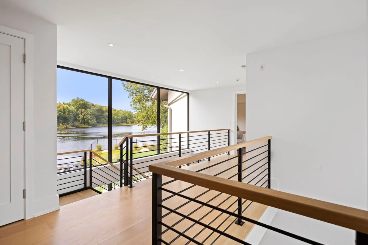Interior of a modern home with a wooden staircase railing, white walls, ceiling lights, and large windows showing a river and trees outside.