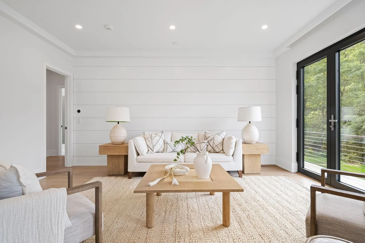 Living room with white walls, large glass sliding door, beige sofa with cushions, wooden coffee table with a vase and decorative object, two side tables with lamps, armchairs, and a natural fiber rug.