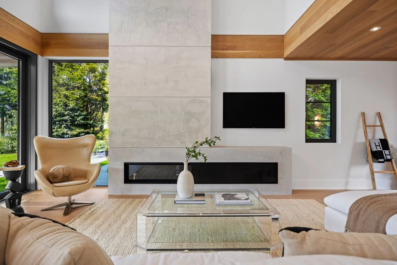Modern living room with large windows showing greenery outside, a wall-mounted TV, a glass coffee table with a white vase and magazines, a beige armchair with a cushion, a woven rug, and a wooden ladder with black and white towels.