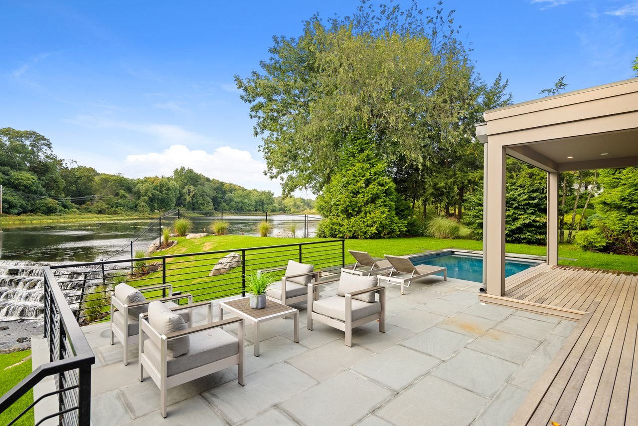 Outdoor patio with white seating and lounge chairs near a small pool, overlooking a grassy yard, a river, and a wooded landscape with a waterfall feature.