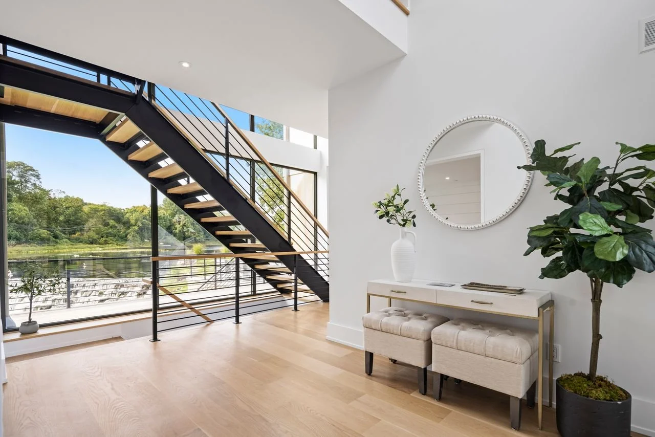 Modern interior space with a staircase, large windows overlooking a river, a white console table with a vase and mirror, and large potted plants.
