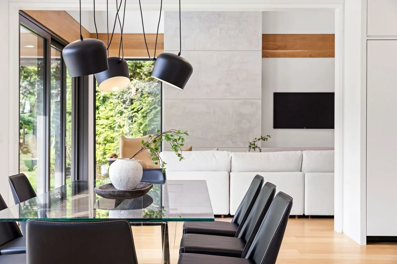 Modern dining and living room with black pendant lights, glass dining table with a vase, black chairs, white sofa, and large windows showing greenery outside.