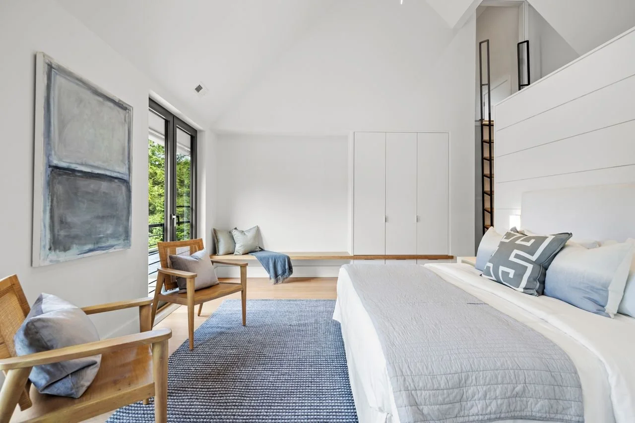 Modern bedroom with white walls, a large bed with decorative pillows, wooden chairs with cushions, a blue and white area rug, a built-in bench with pillows, and a large window with a balcony view.