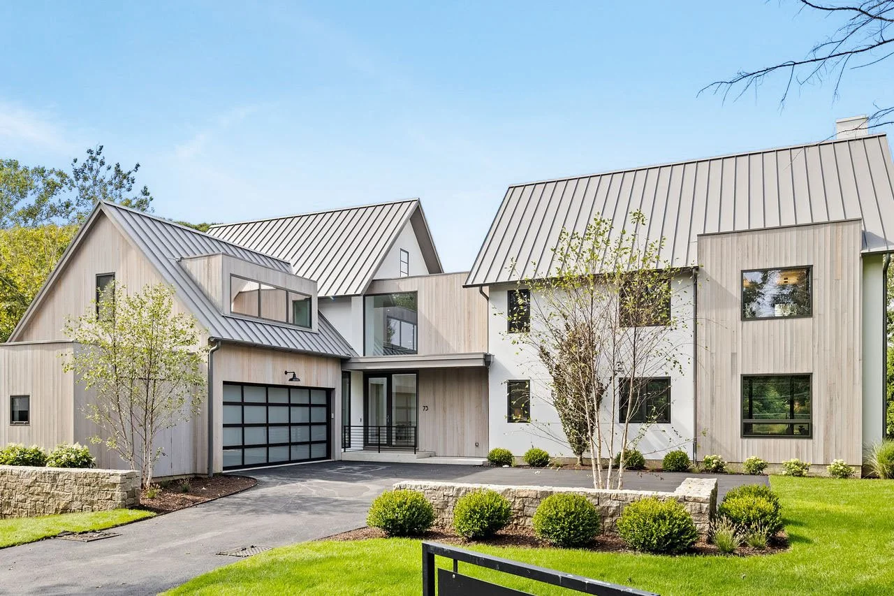 Modern multi-story house with a metal roof, surrounded by green lawn and trees, under a clear blue sky.