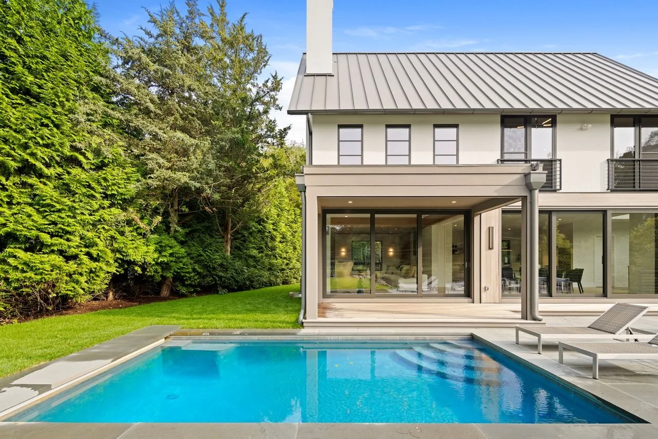 Modern house with large glass doors, gray roof, and two lounge chairs beside a swimming pool, surrounded by a green lawn and tall trees.