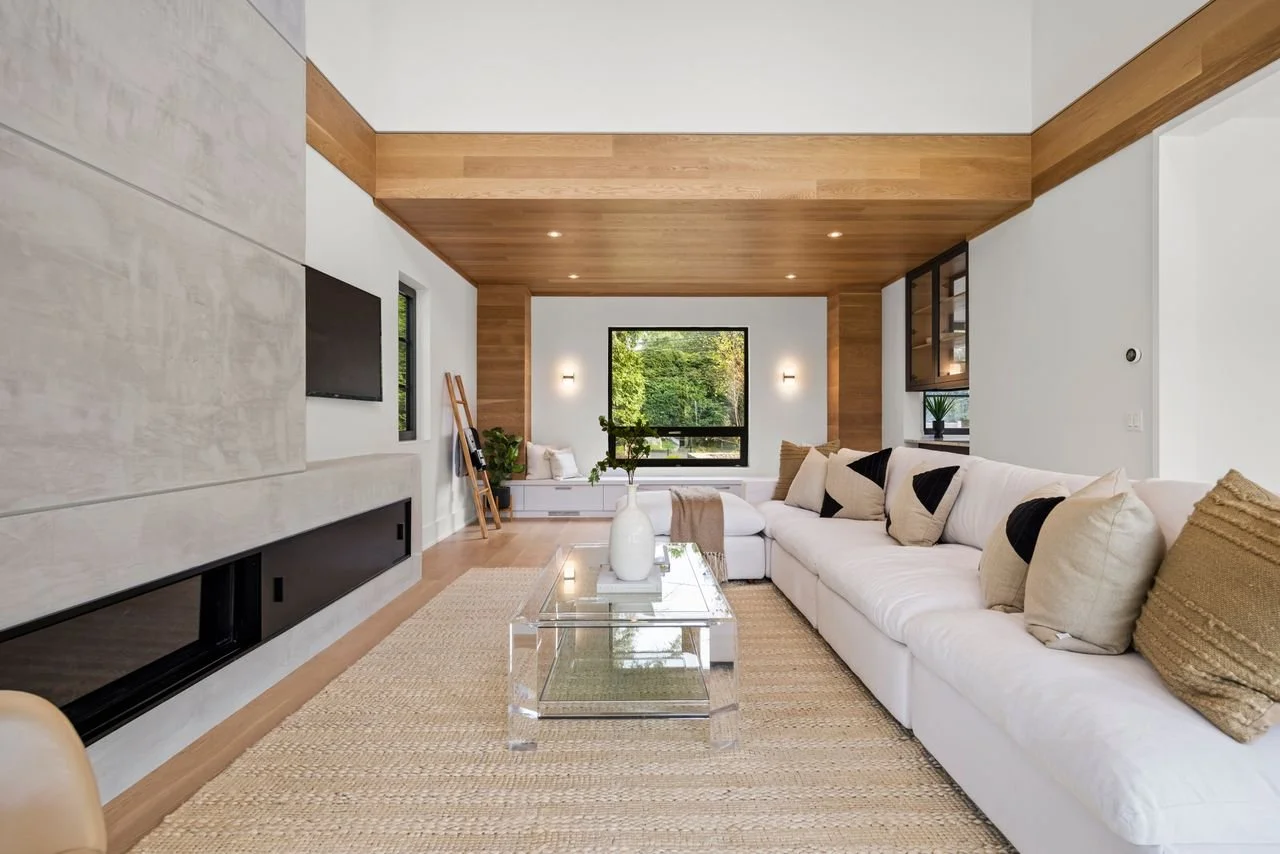 Modern living room with white sectional sofa, throw pillows, glass coffee table, and large window overlooking greenery.