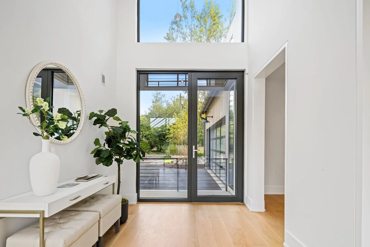 Modern interior entryway with white walls, a sliding glass door leading to an outdoor deck, and a large window above the door letting in natural light. Decor includes a white vase with flowers, a mirror, a potted plant, and a beige bench.