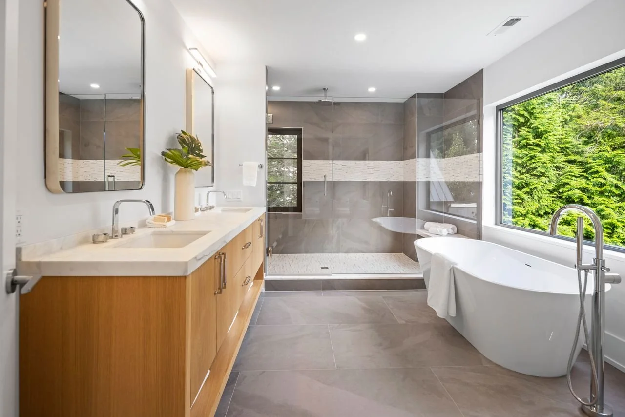 Modern bathroom with a large window, freestanding bathtub near the window, double sink vanity with mirrors, gray tiled walk-in shower with glass doors, neutral color palette, greenery outside.