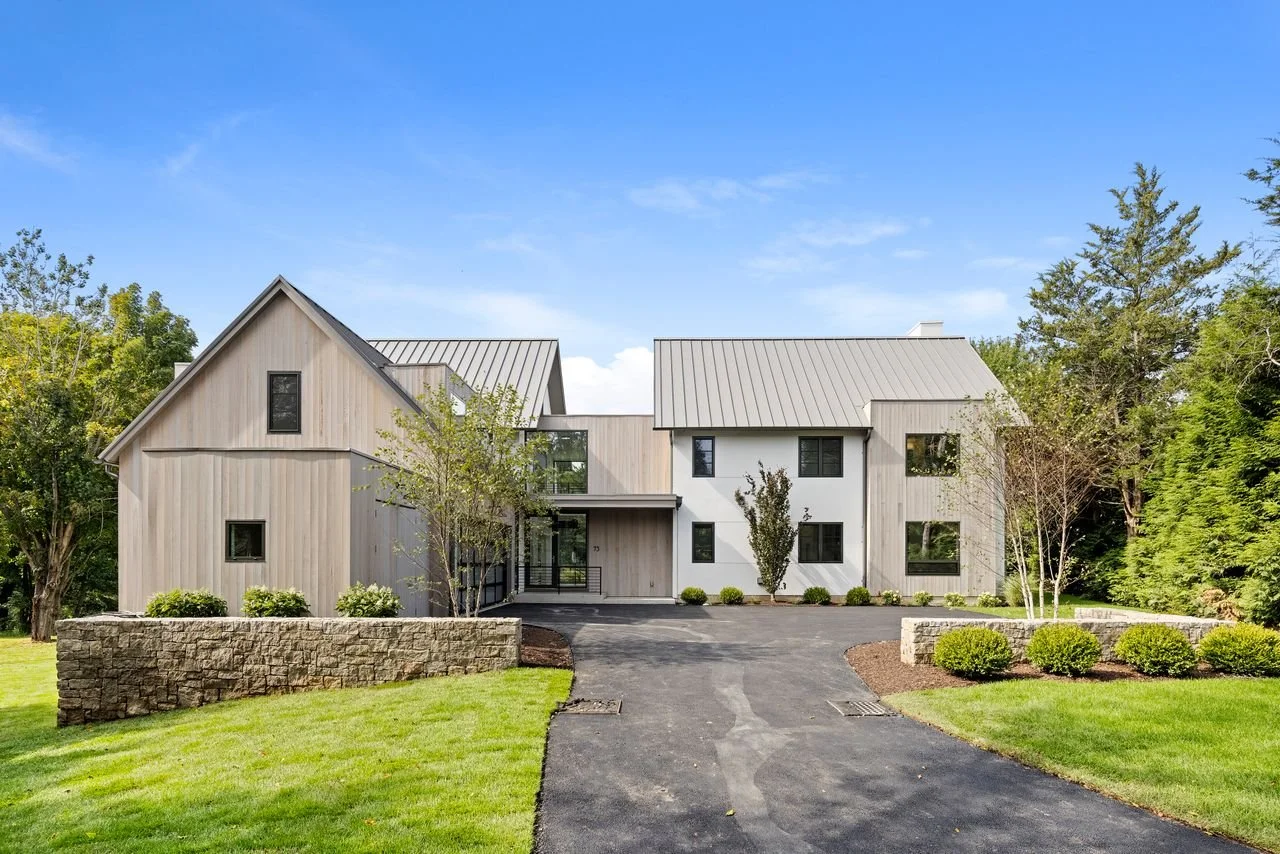 Modern two-story house with a gray metal roof, light wood and white exterior walls, surrounded by green trees and grass, with a paved driveway leading to the front entrance.