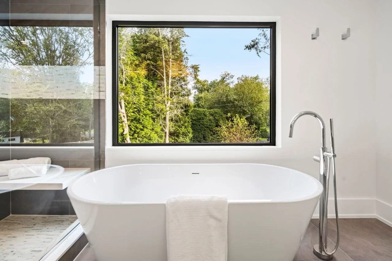 Modern bathroom with a white bathtub, large window showing a view of trees, and a glass shower enclosure.