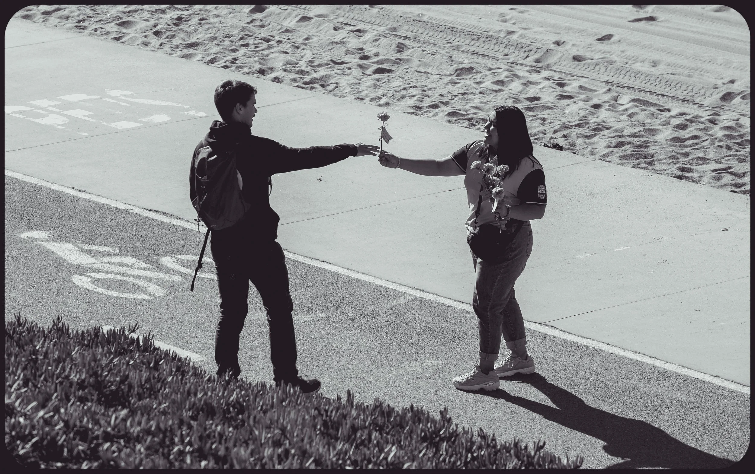 A young man is handing a woman flowers on a sidewalk near a beach, with the woman smiling and requesting the flowers.