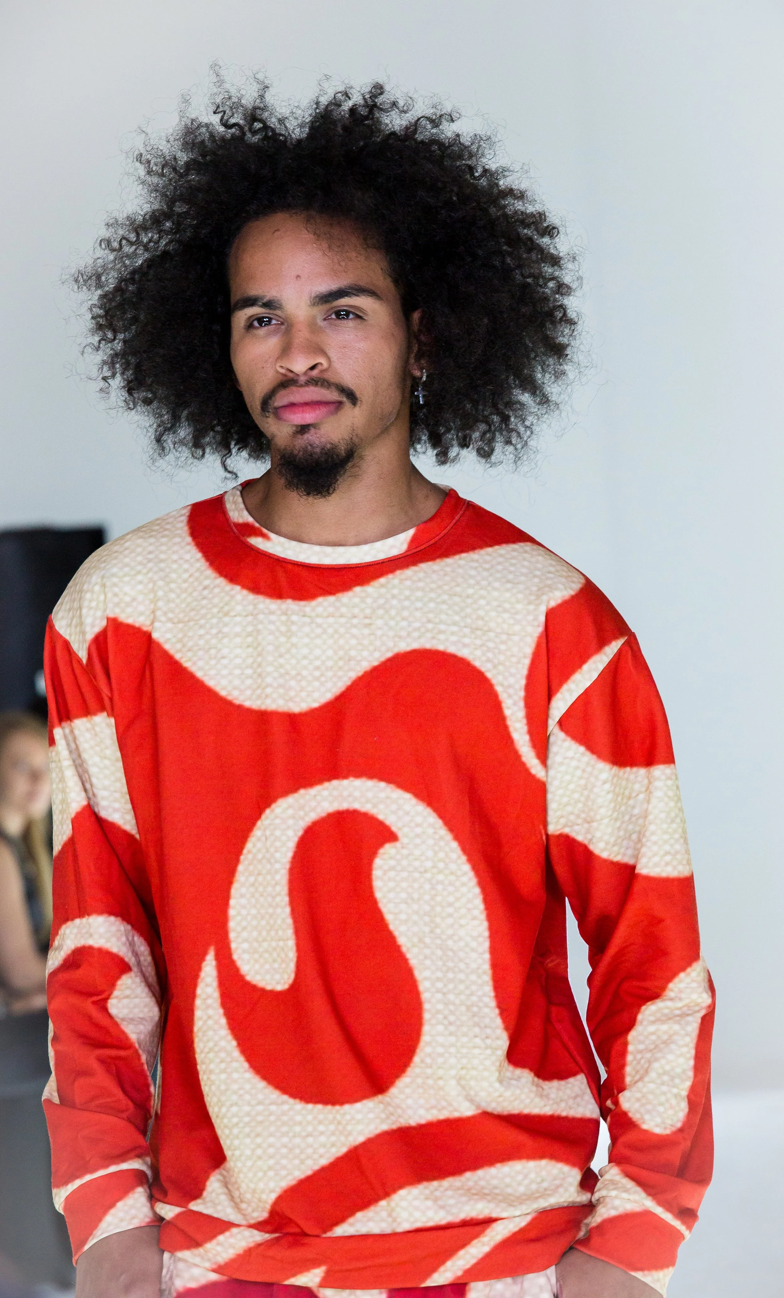 A man with curly black hair, a mustache, and a goatee wearing a red and cream patterned long-sleeve shirt, standing indoors with a neutral expression.
