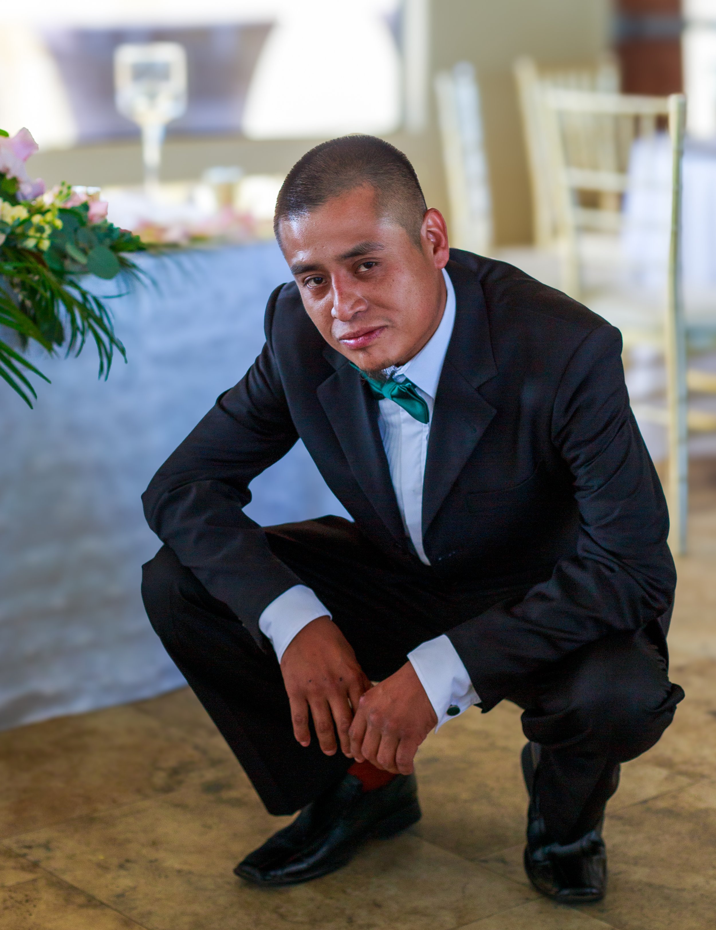 A man in a black suit and green bow tie crouching on the floor in a banquet hall, looking at the camera.