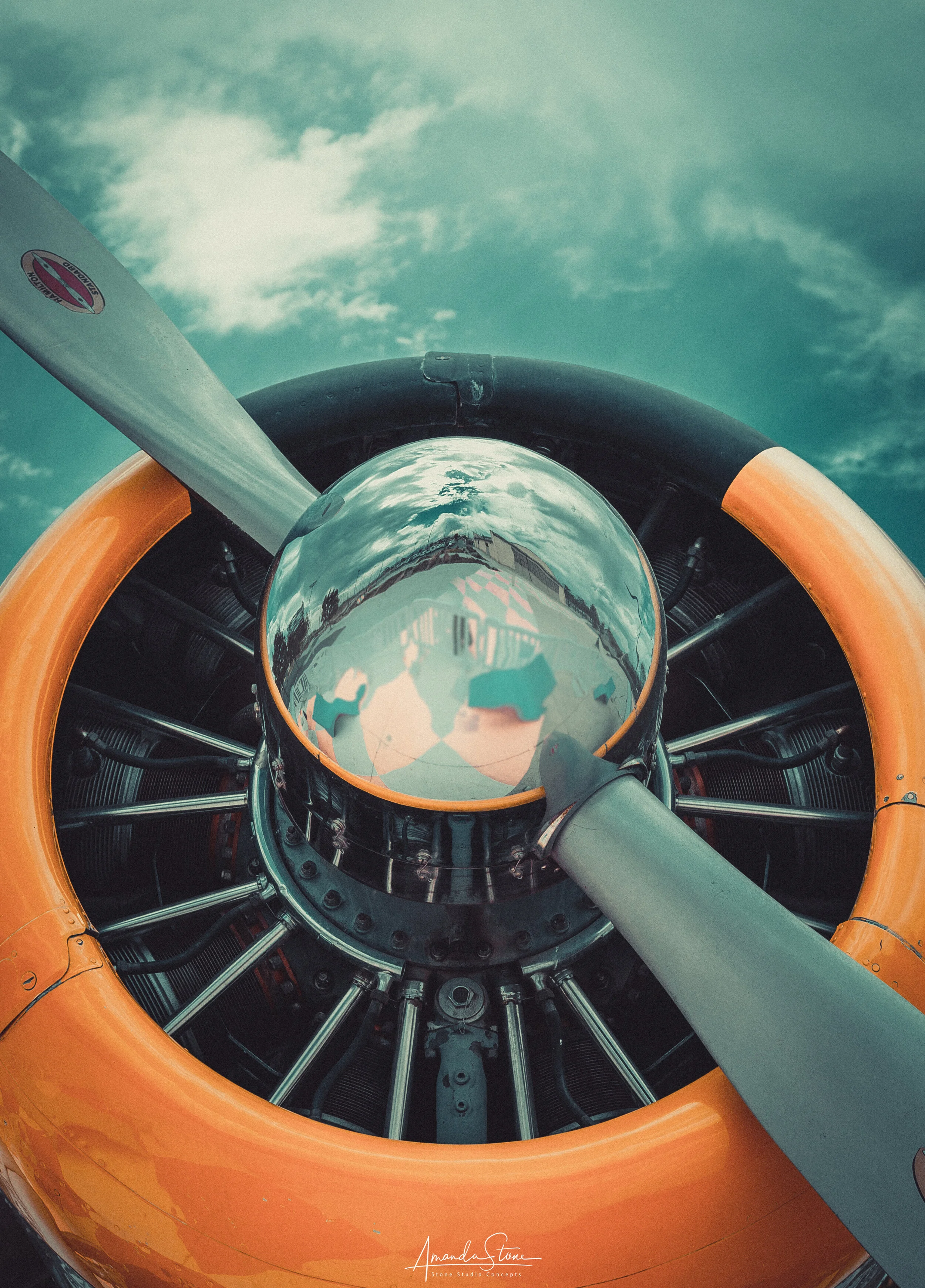 Close-up of a vintage aircraft engine with a reflective glass nose cone, propeller blades, and a partially visible bright orange cowling against a cloudy sky.