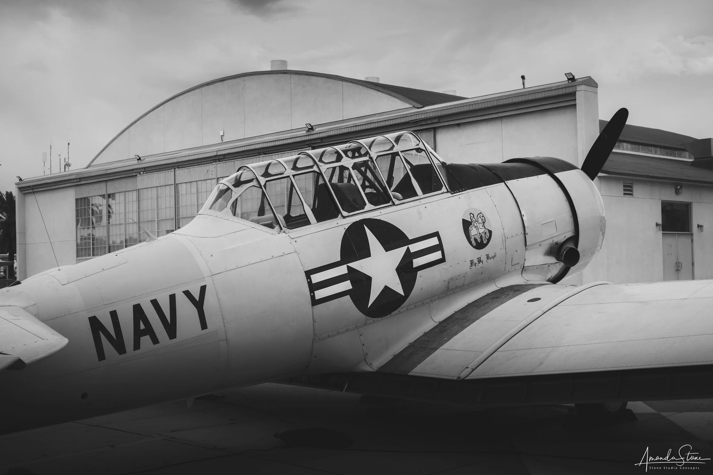 Black and white photo of a vintage Navy aircraft with a glass cockpit and a large propeller, parked outside a hangar.