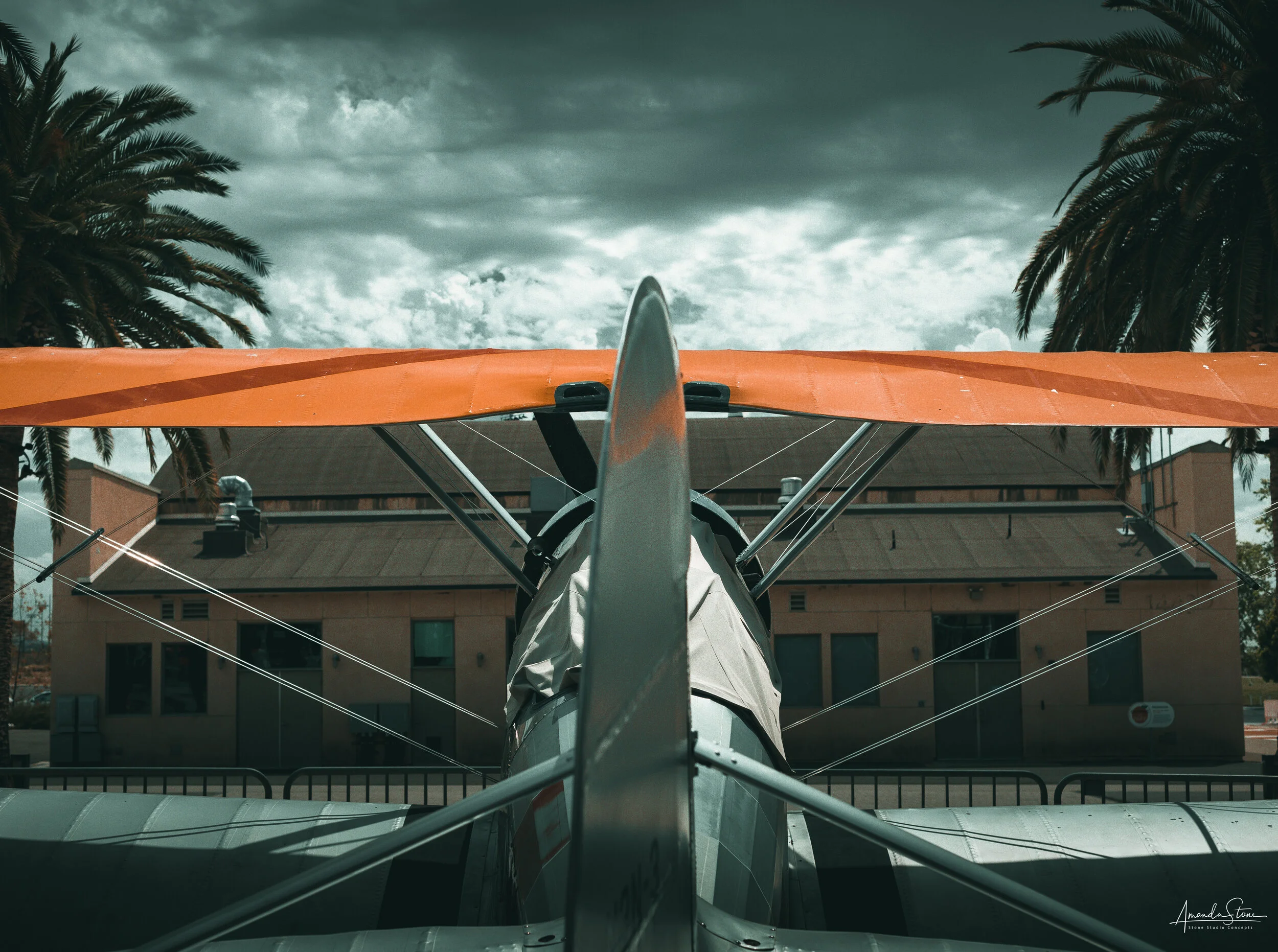 Front view of a small airplane with an orange wing, parked in front of a building with palm trees and a cloudy sky in the background.