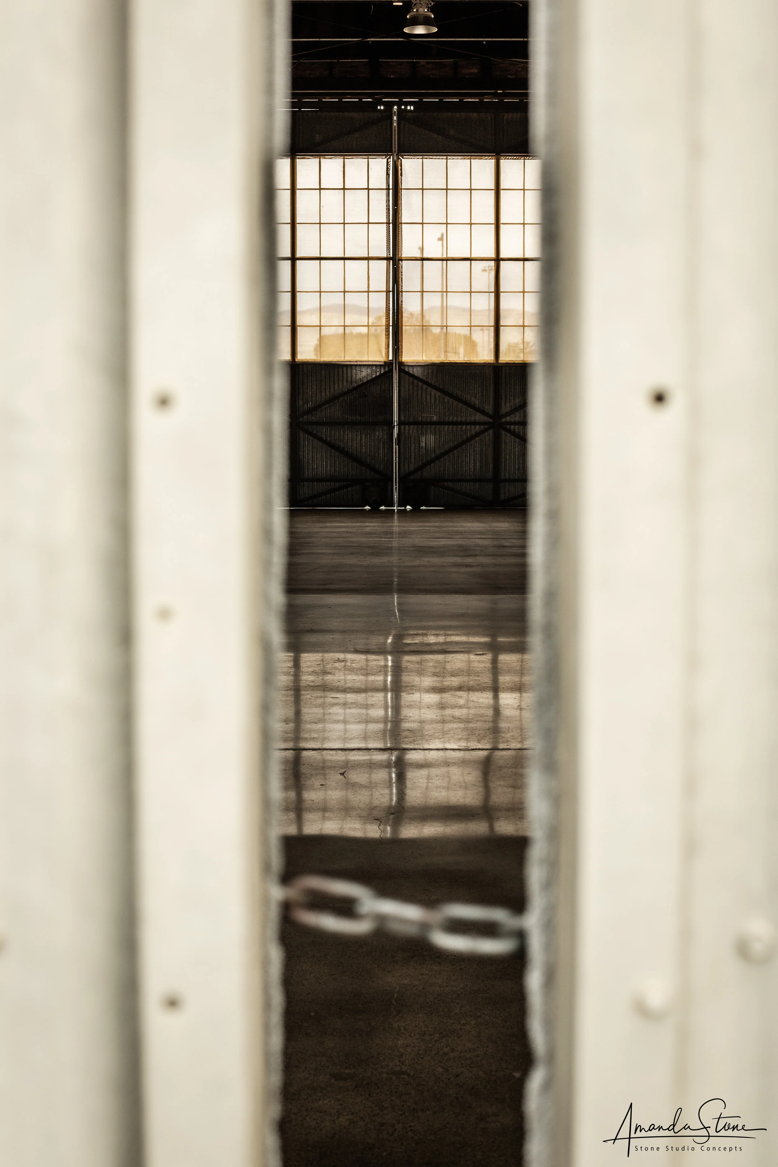 View through a narrow gap in a metal door revealing an industrial interior with chain-link fence and a large window on the far wall, allowing natural light to enter.