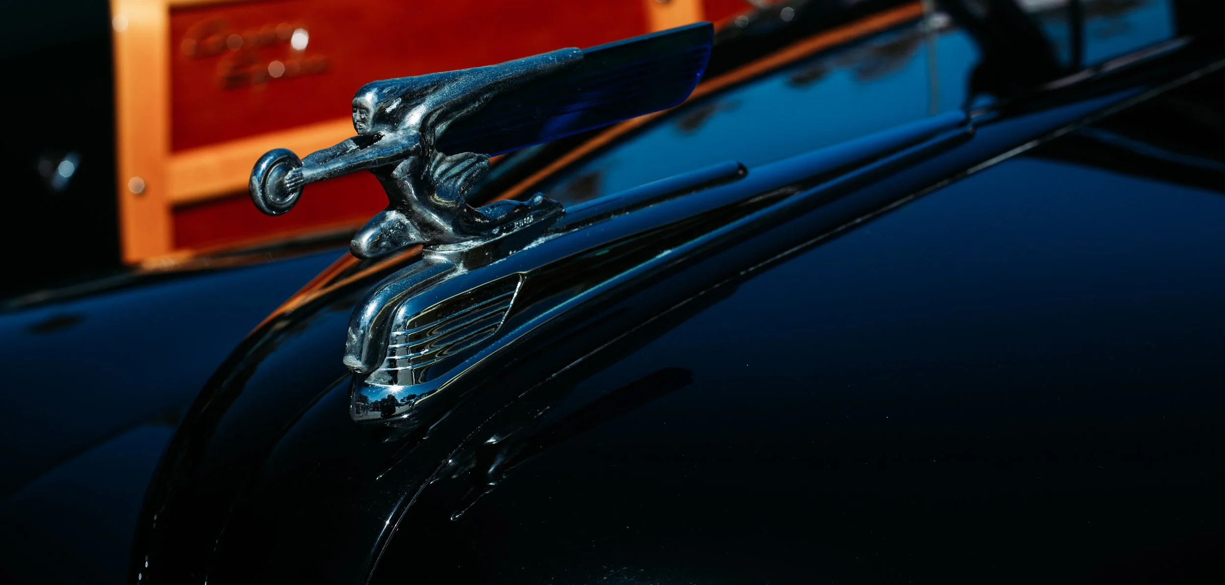 Close-up of a shiny vintage car hood ornament in the shape of a woman with outstretched arms, mounted on a black car.