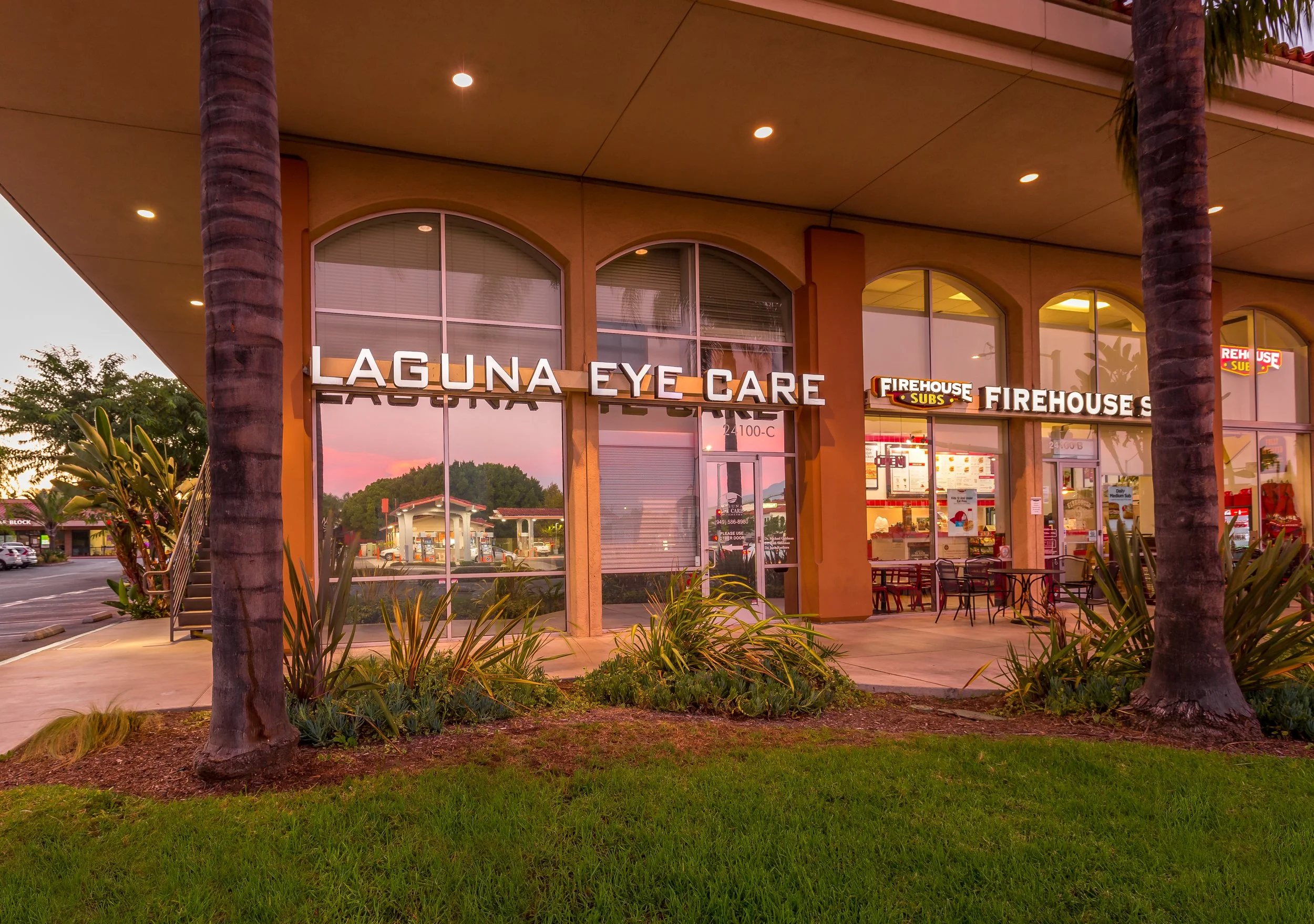 A strip mall with large glass windows, palm trees, and green grass. The stores include Laguna Eye Care and Firehouse Subs, with the signs visible above the entrances. The scene is at sunset.