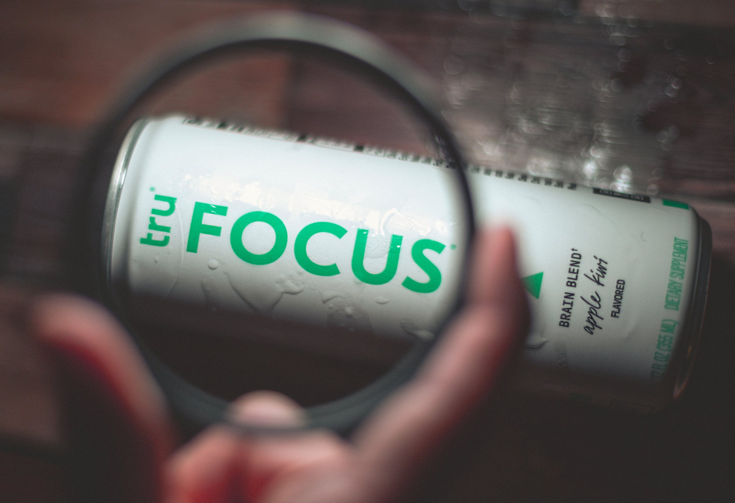 Close-up of a can of Tru Focus brain blend apple cinnamon flavored beverage. The can is viewed through a magnifying glass, which enlarges and highlights the word 'FOCUS' on the can.