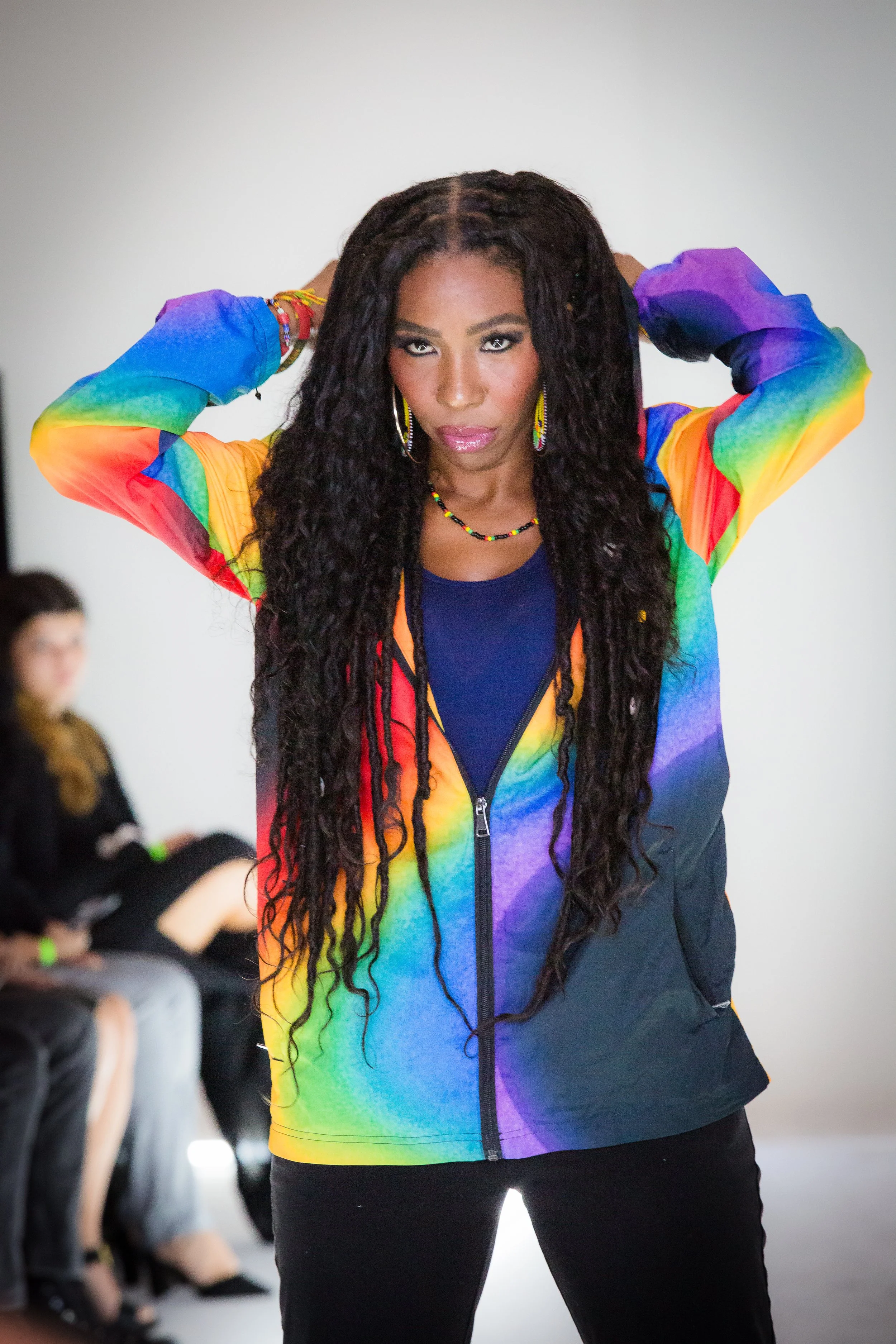 Woman with long curly hair wearing a colorful rainbow jacket posing with hands behind her head, in front of a plain background.