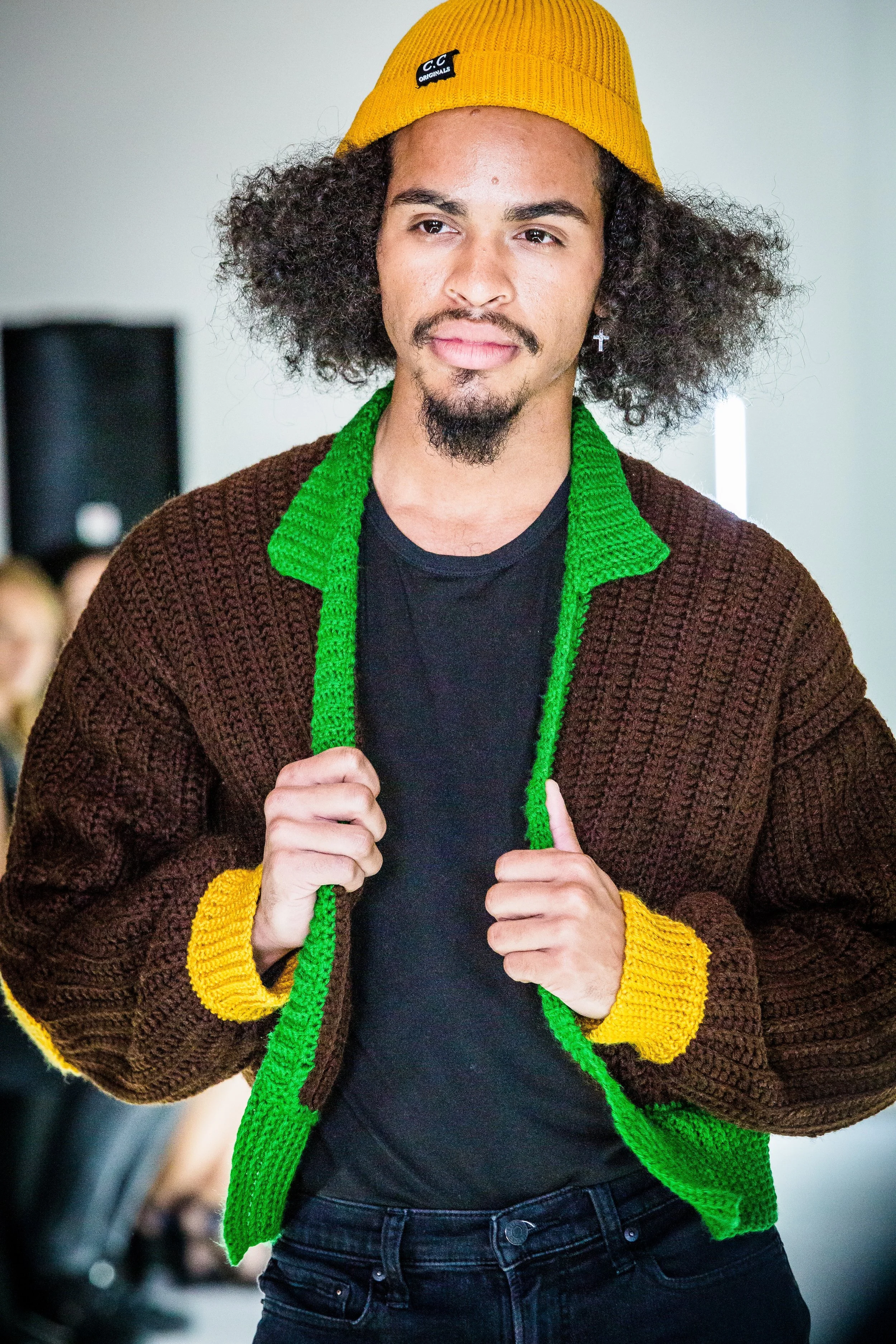 A man modeling in a colorful, chunky-knit sweater with green accents, yellow cuffs, and a brown body, paired with a yellow beanie with a black and white logo. The man has curly hair, a mustache and goatee, and is holding the collar of his sweater wit