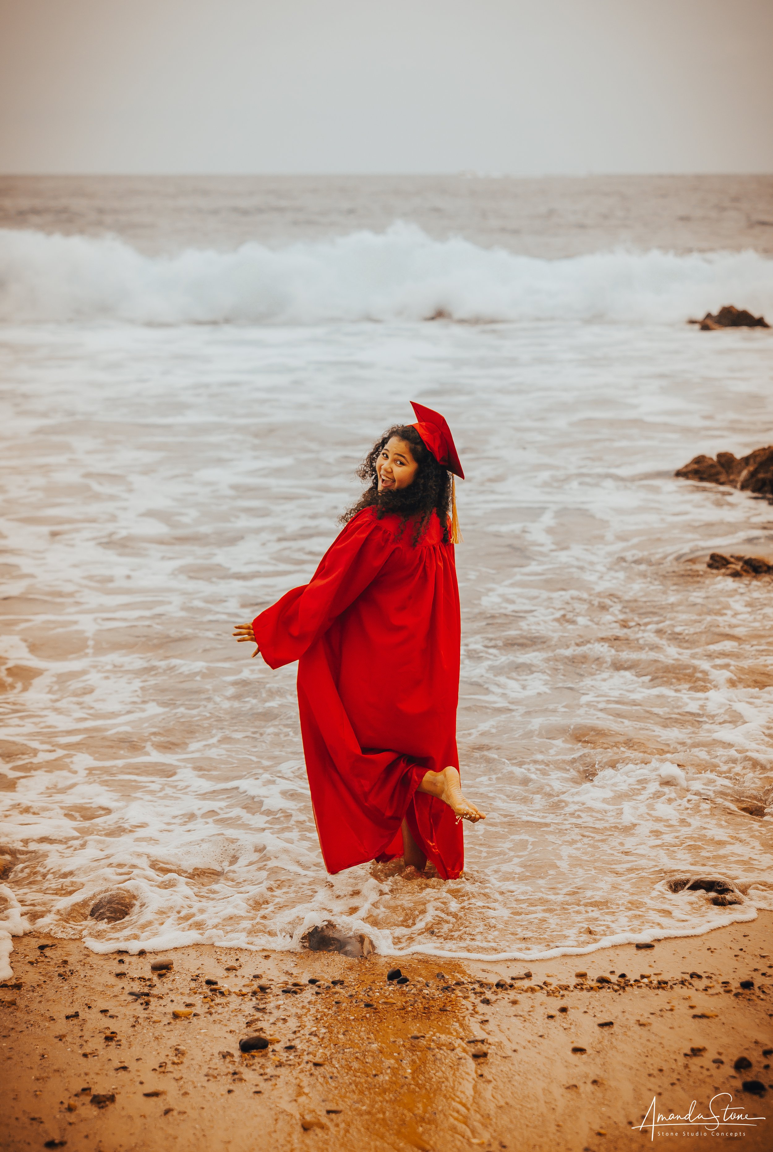 Young woman in red graduation gown and cap standing in the ocean with waves crashing around her, smiling at the camera.