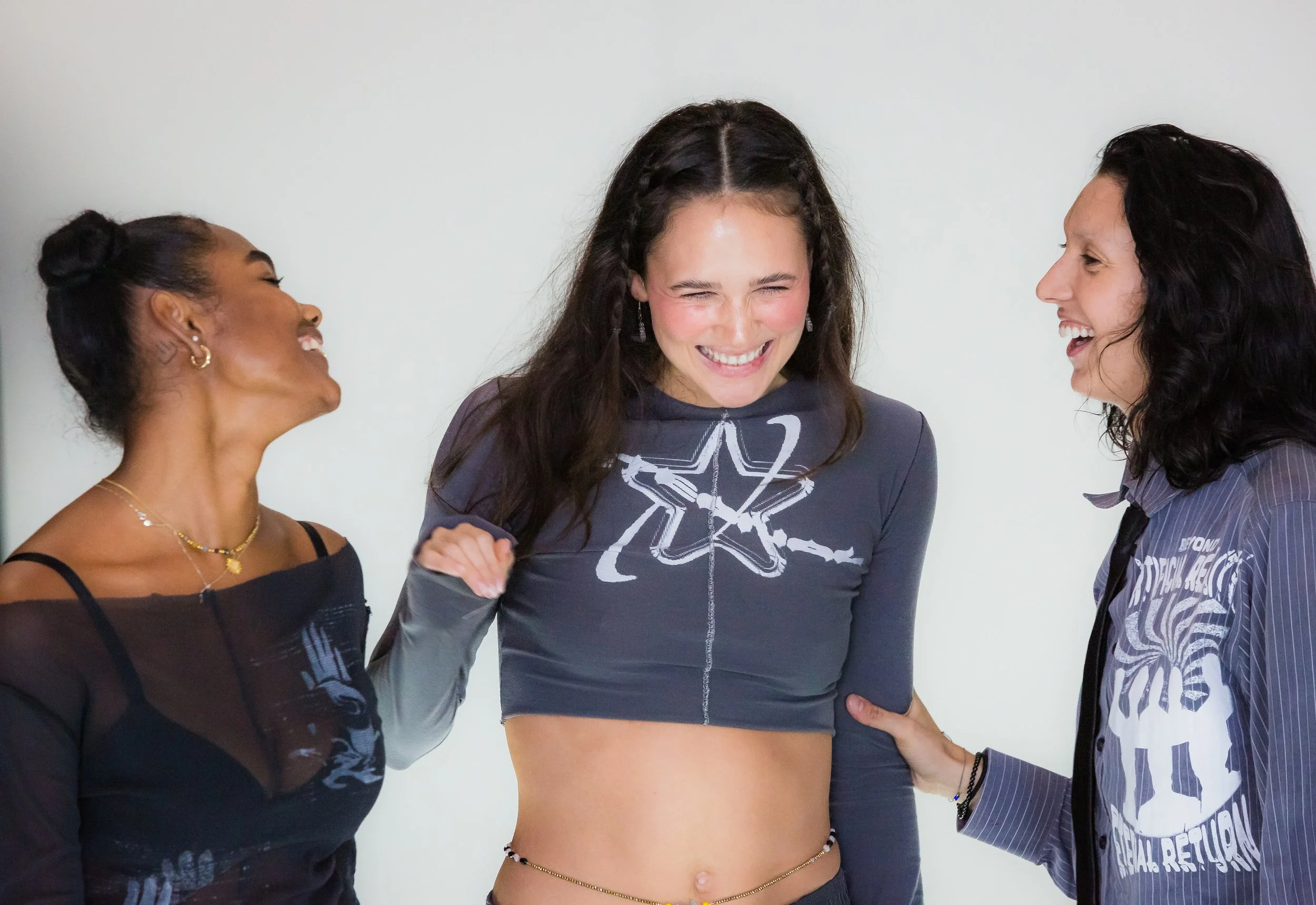 Three women laughing and smiling together in casual clothing, standing against a plain background.