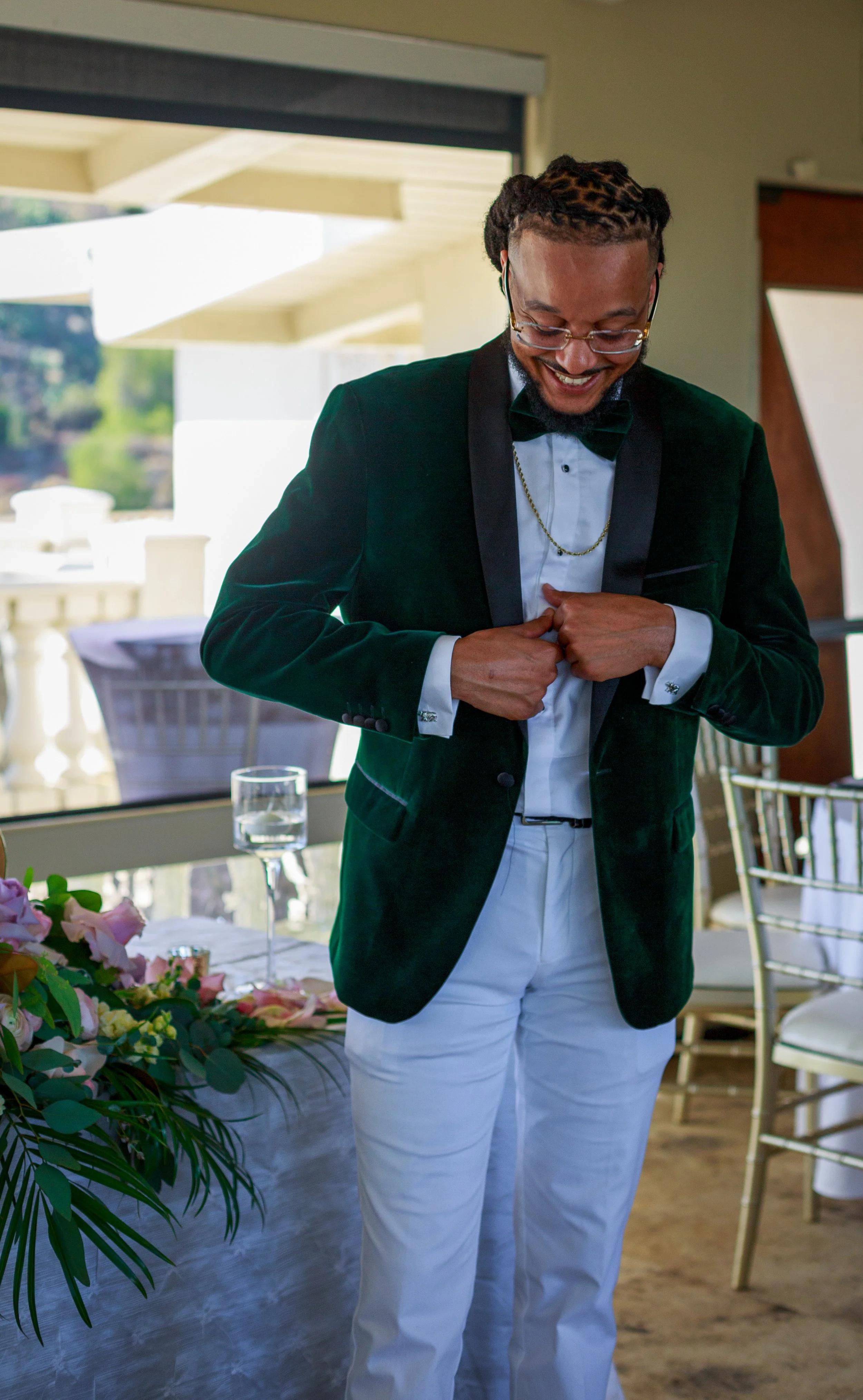 Man smiling, adjusting tuxedo jacket, wearing glasses, black tuxedo with a bow tie, white shirt, white pants, gold chain, in a decorated room for a celebration.