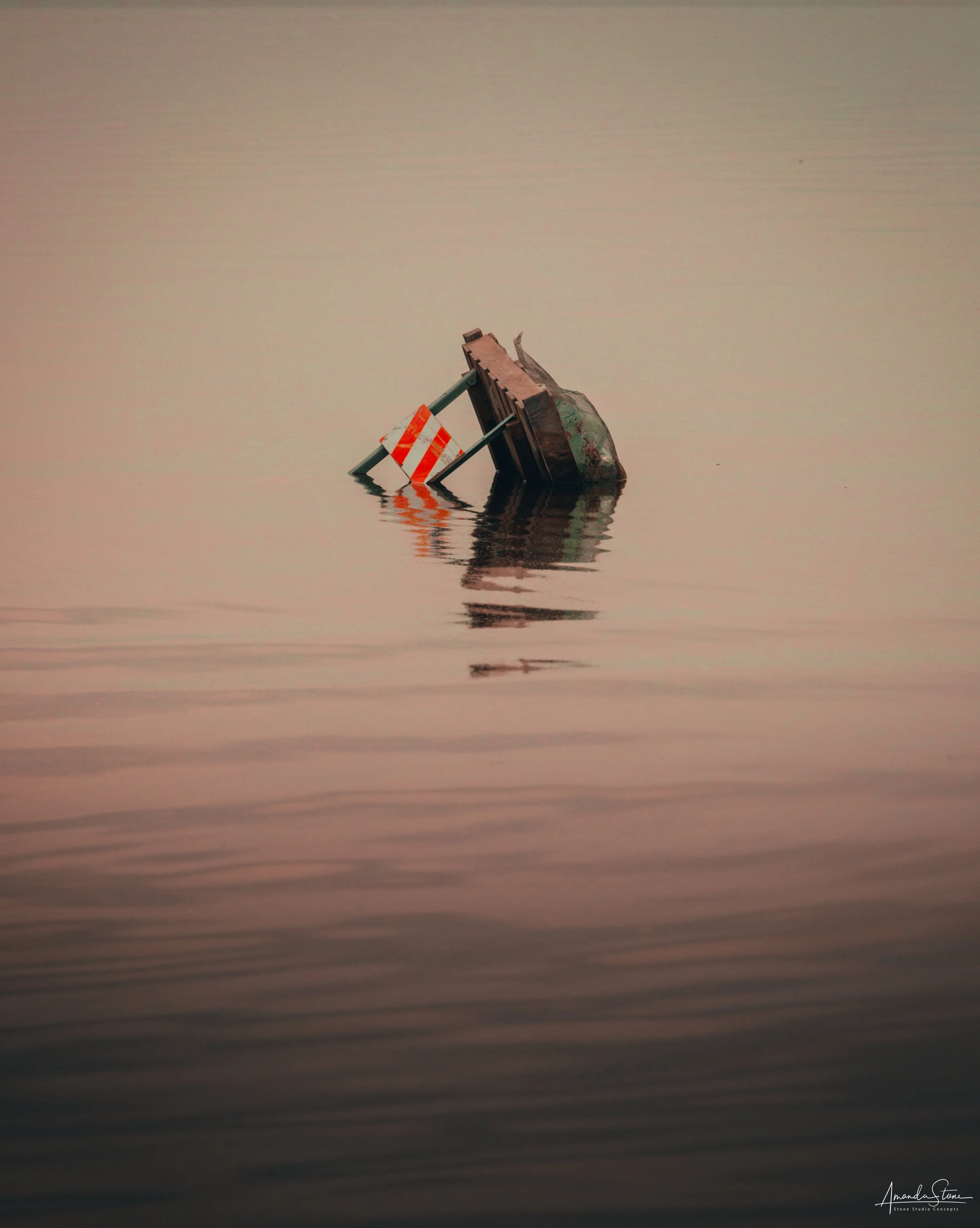 A partially submerged road barrier and a metal barrier with orange and white stripes in a body of water, with gentle ripples and a hazy, pinkish sky.