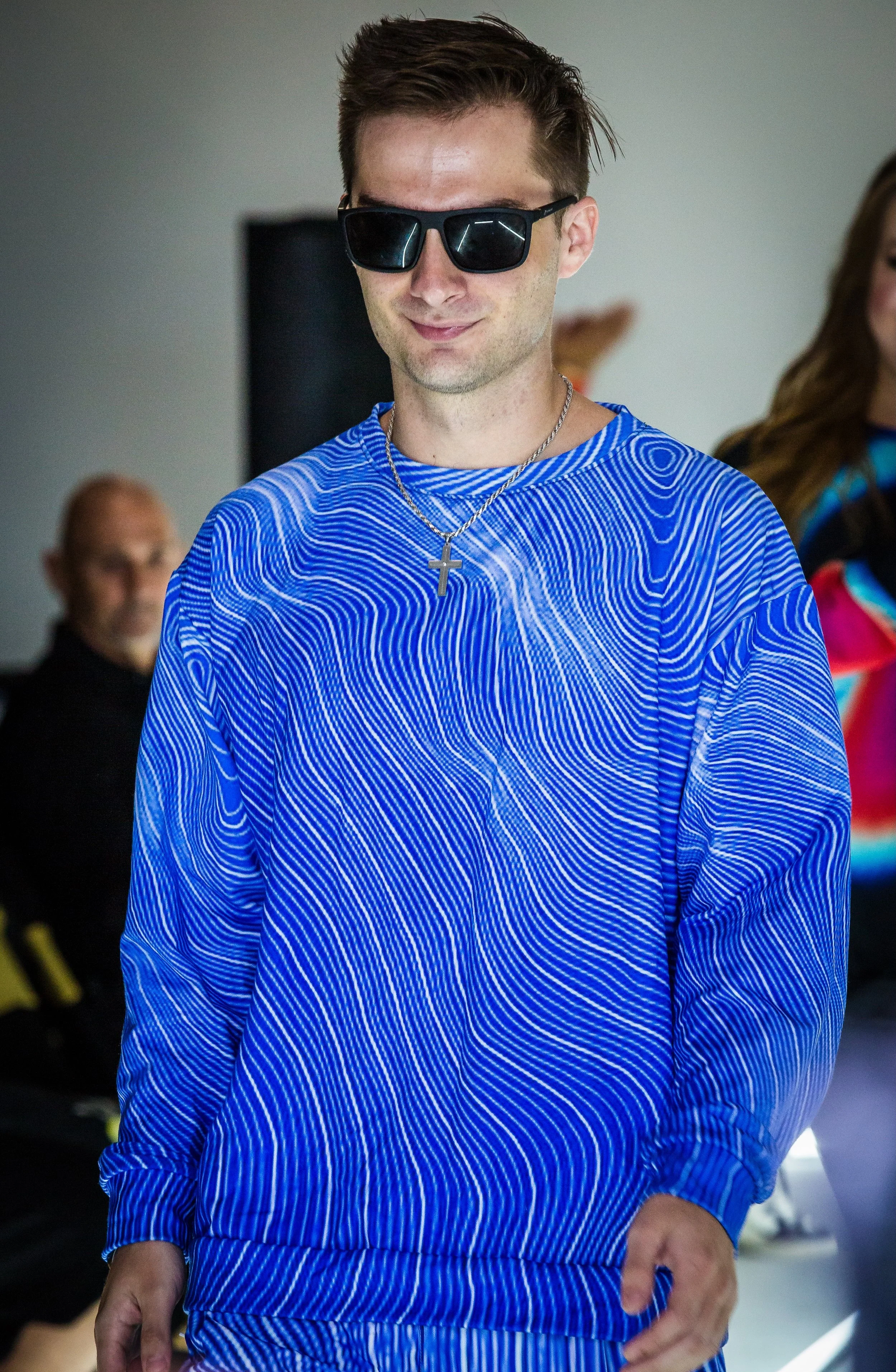 A young man with light skin, wearing dark sunglasses, a blue patterned sweatshirt, and a silver cross necklace. He is smiling slightly and standing indoors with other people in the background.