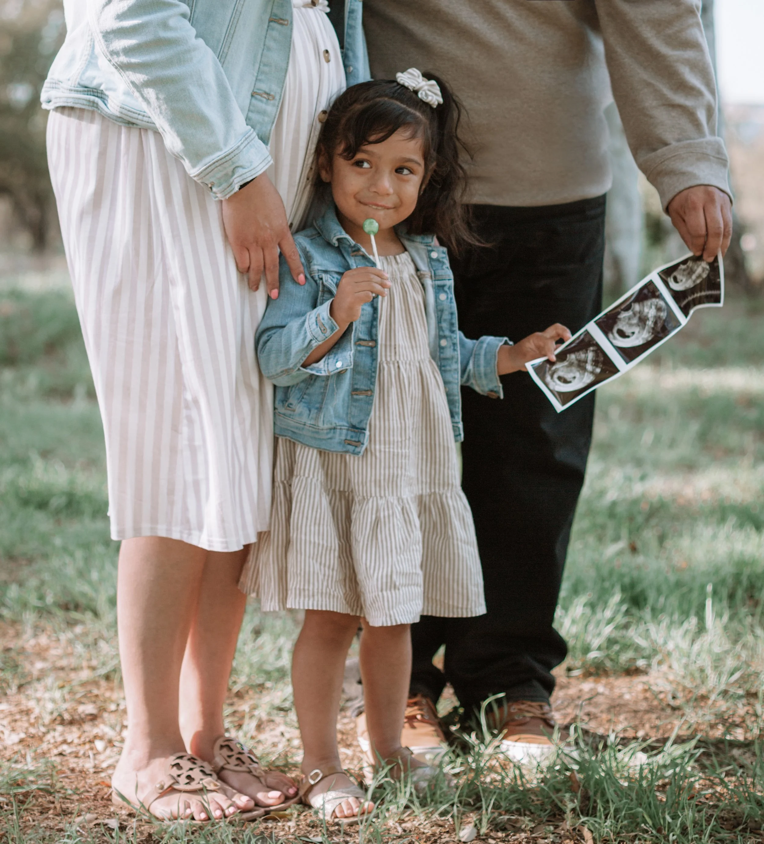 A young girl holding a lollipop and an ultrasound image, standing between two adults outdoors.