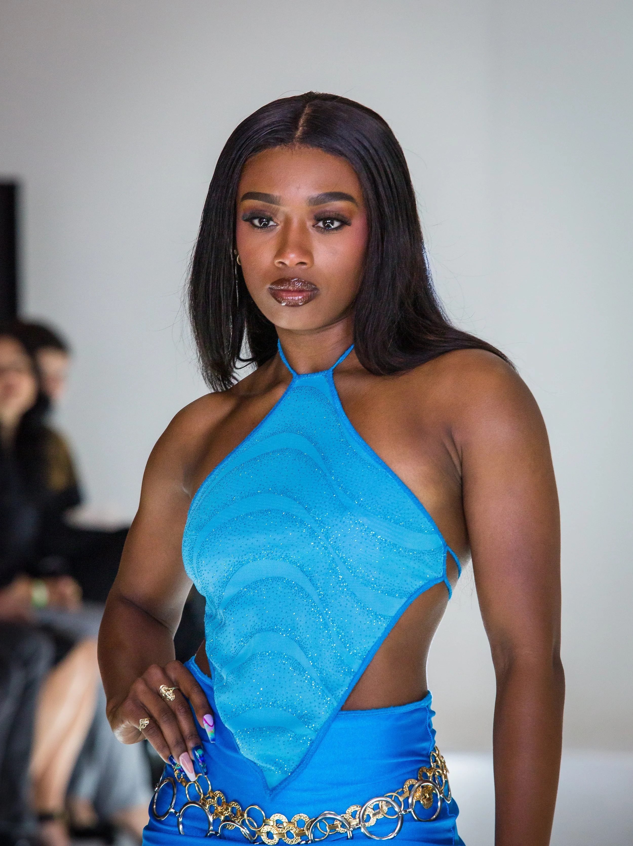 A female model walking on a runway wearing a vibrant blue halter top with cutouts and embellishments, paired with a matching belt and blue skirt, during a fashion show.