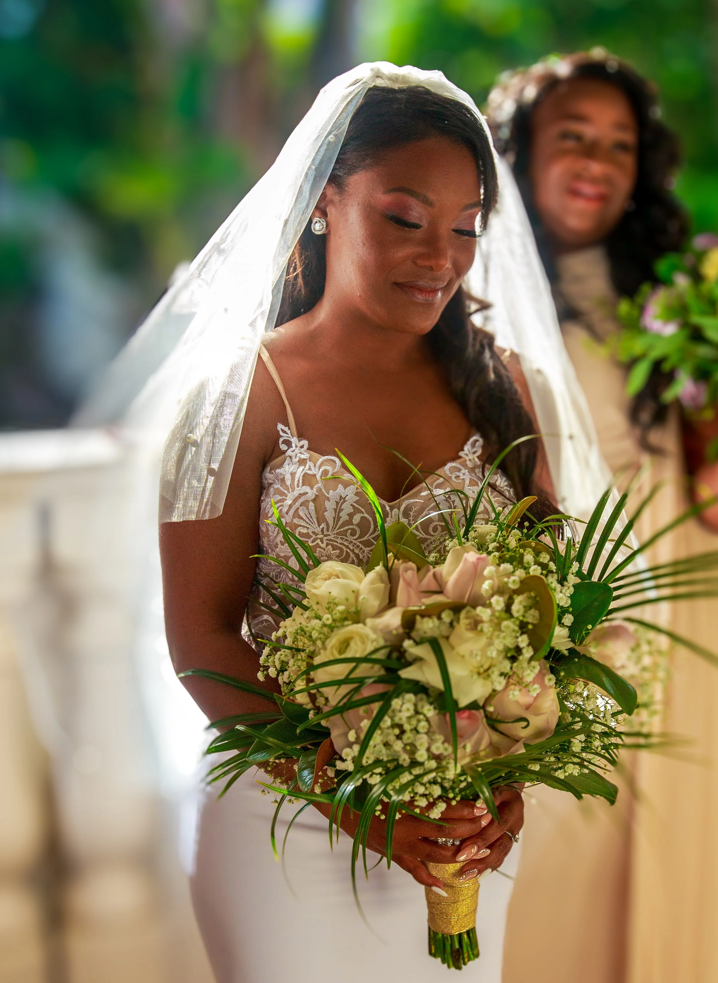 A bride in a white wedding dress holding a bouquet of white and pink roses with greenery, standing in a garden. Another woman in a beige dress and floral headband is in the background, holding a bouquet.