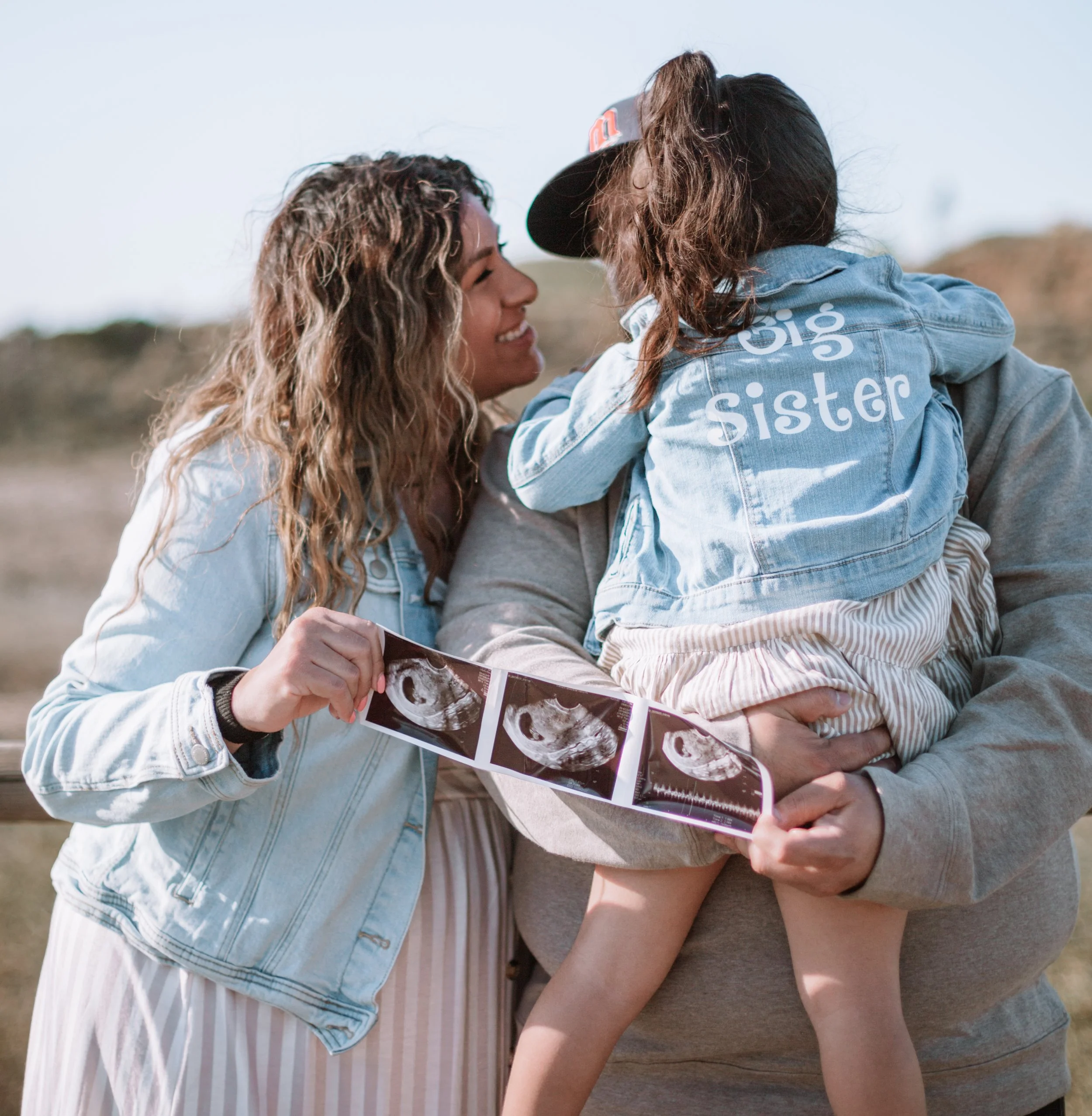 A woman and a man holding a young girl, who is wearing a denim jacket with "big sister" written on the back, while looking at ultrasound images outdoors.