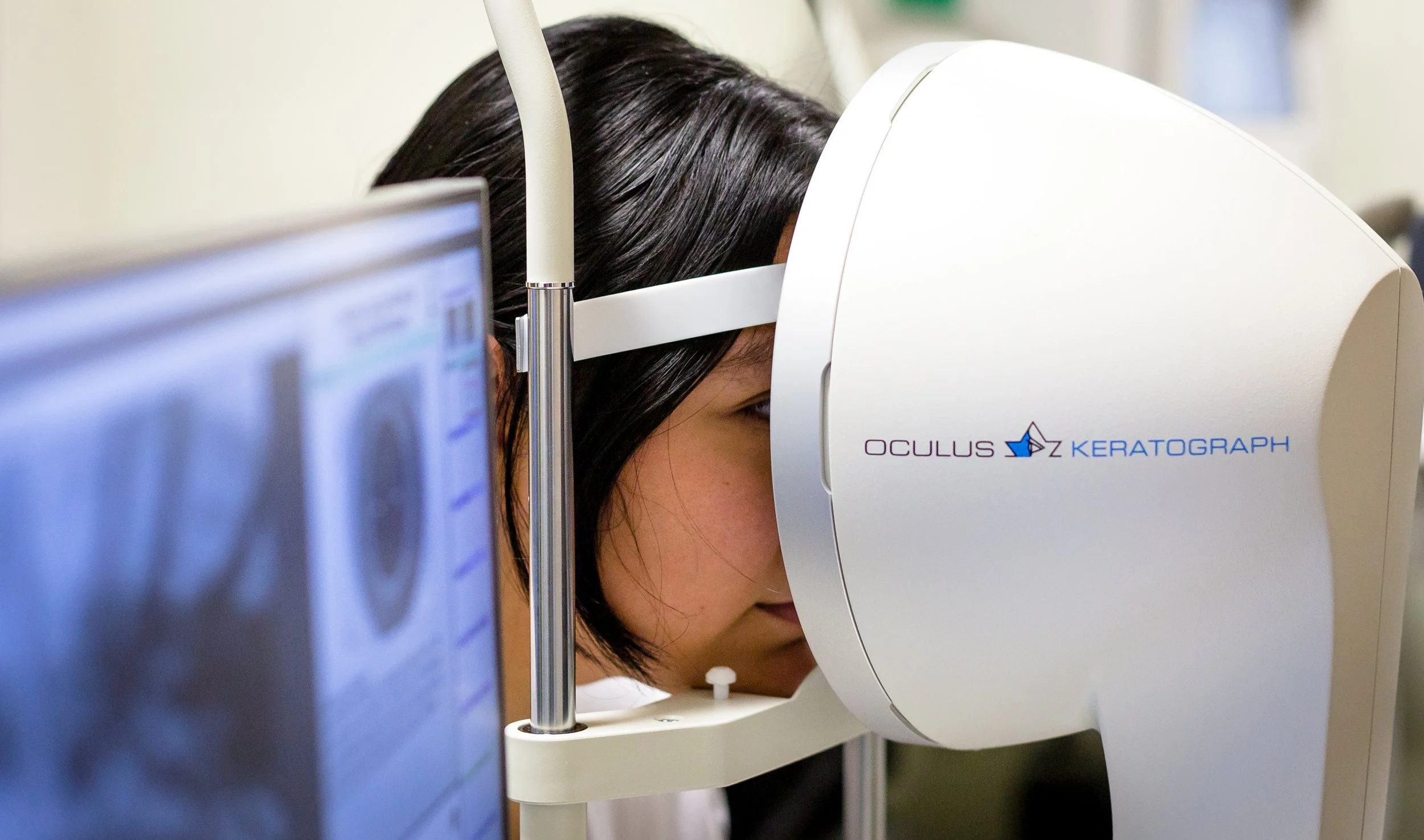 A person having a laser eye procedure using an Oculus Keratograph device, with a computer screen displaying eye scans nearby.