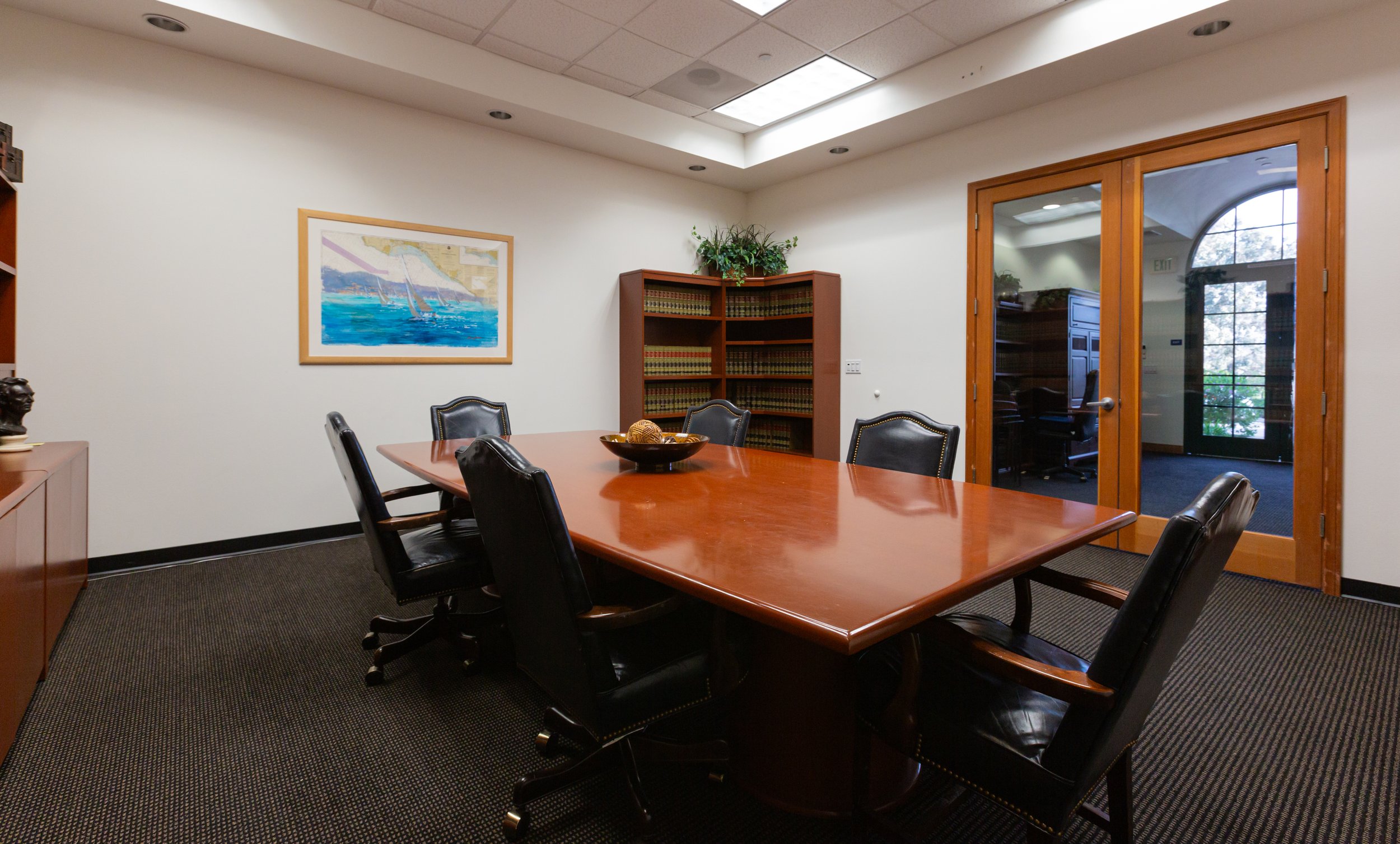 Conference room with a large wooden table, six black leather chairs, a painting of sailboats, a bookshelf with legal books, a bowl with decorative balls, a wooden cabinet, and glass double doors leading to a sunlit office.