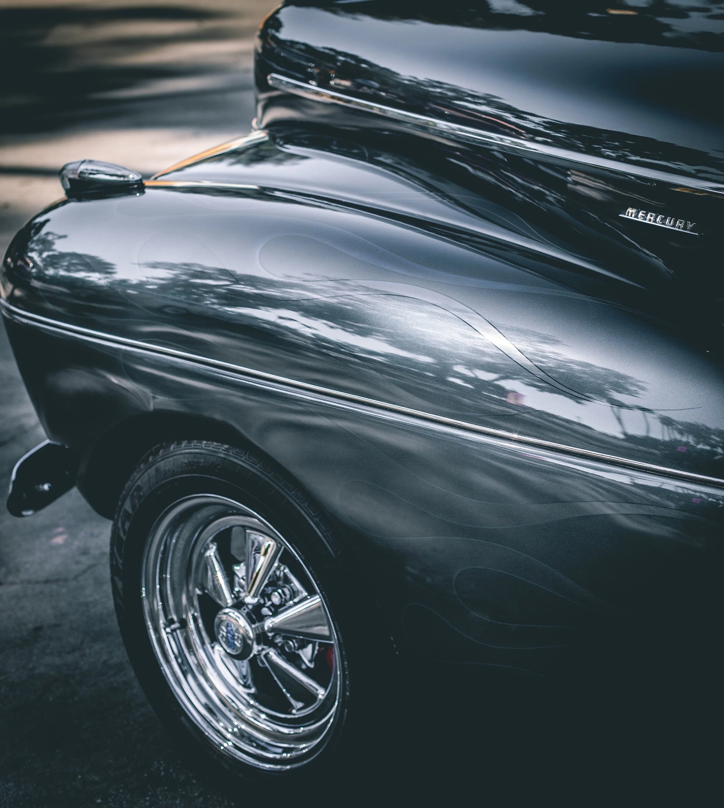 Close-up of a vintage black Mercury car showing the front wheel, fender, and part of the hood, with reflections of trees and sky on the car's shiny surface.