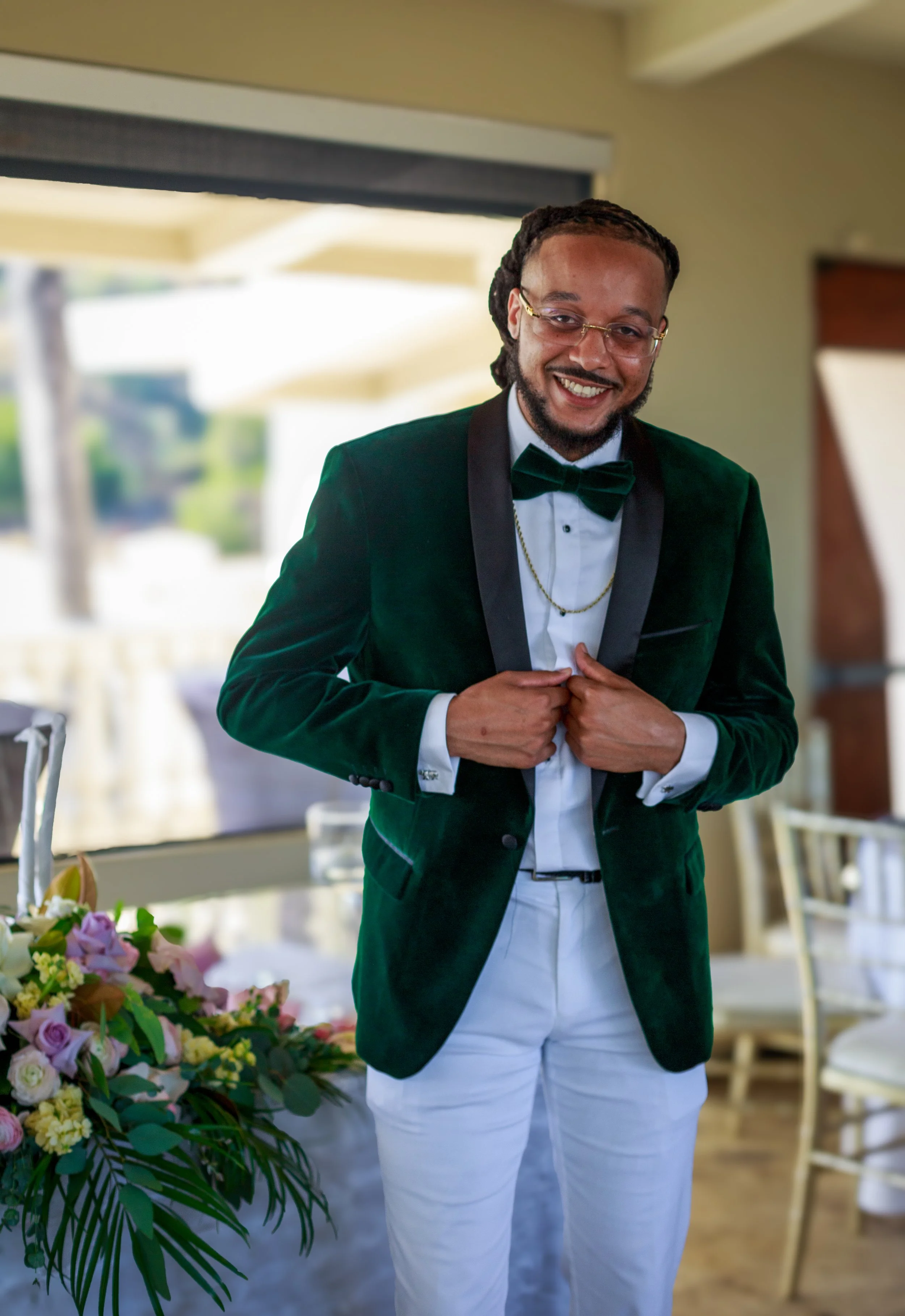 A man dressed in a formal dark green velvet blazer, white shirt, black bow tie, and white pants, standing indoors at a celebration or formal event, smiling and adjusting his blazer.