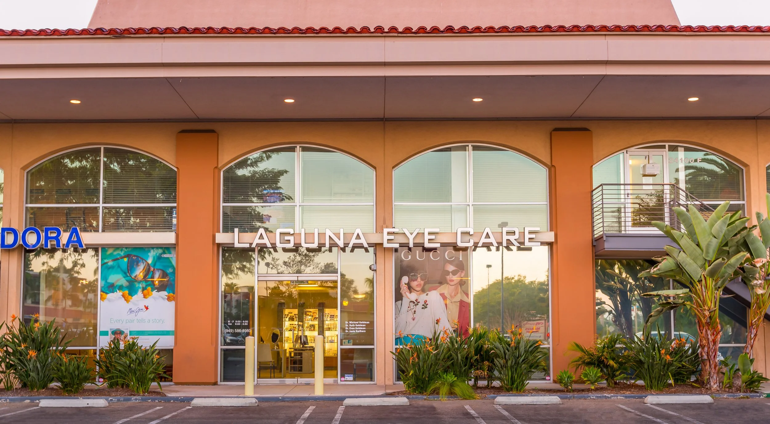 Storefront with large windows, orange pillars, and signage for Laguna Eye Care, Gucci, and Pandora, with plants and parking lot in front.