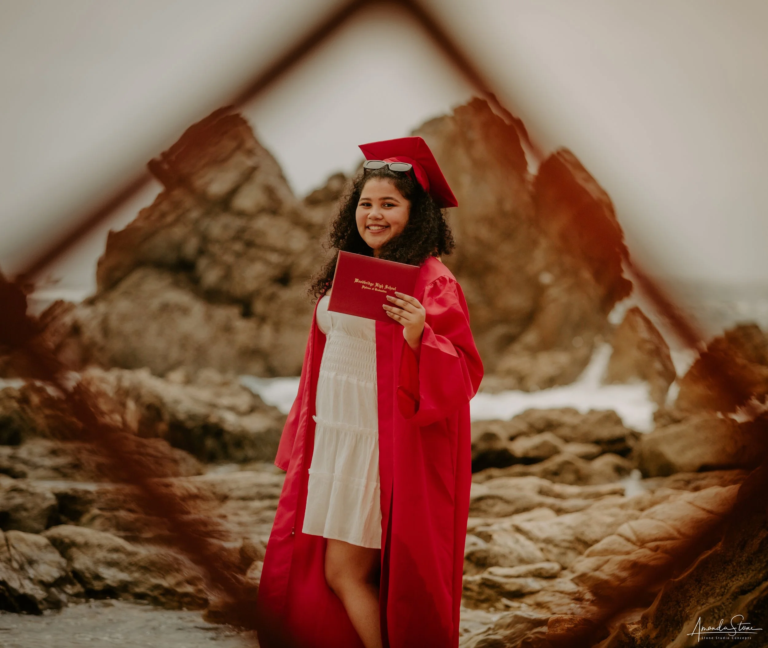 A young woman in a red graduation gown and cap, holding a red diploma, standing on a rocky beach with large rocks in the background, smiling at the camera.