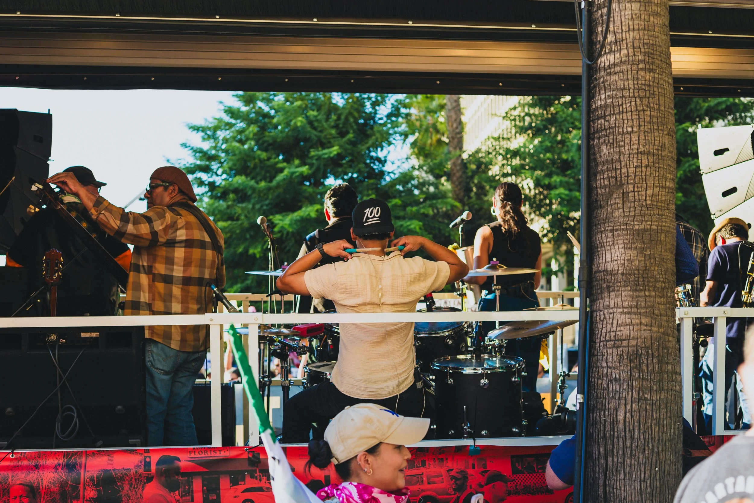 Musicians preparing for a performance on a stage outdoors during the daytime, with trees and buildings in the background.