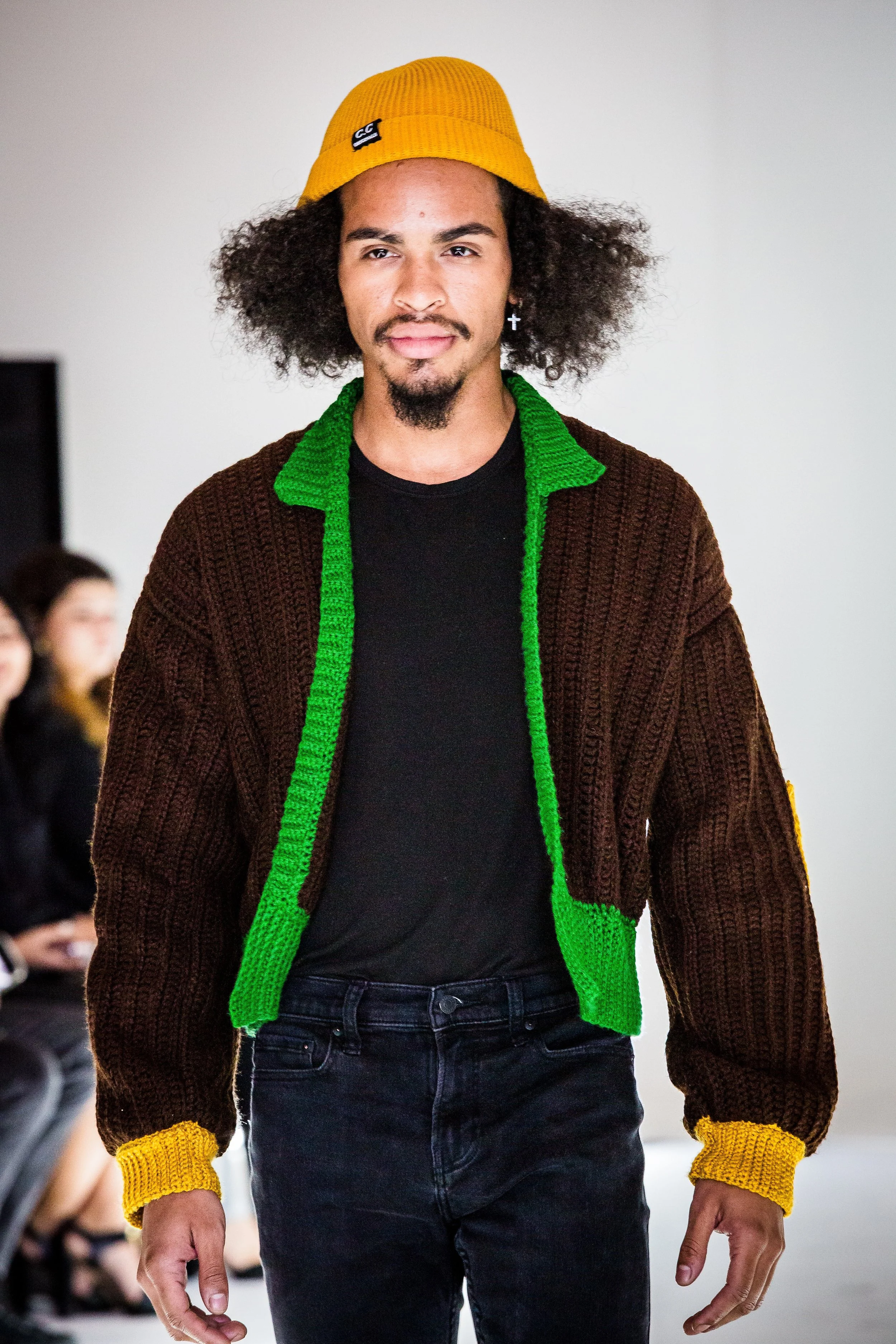 A man with an afro hairstyle, wearing a yellow beanie, a brown sweater with green and yellow accents, and black jeans, walking in a fashion show runway.