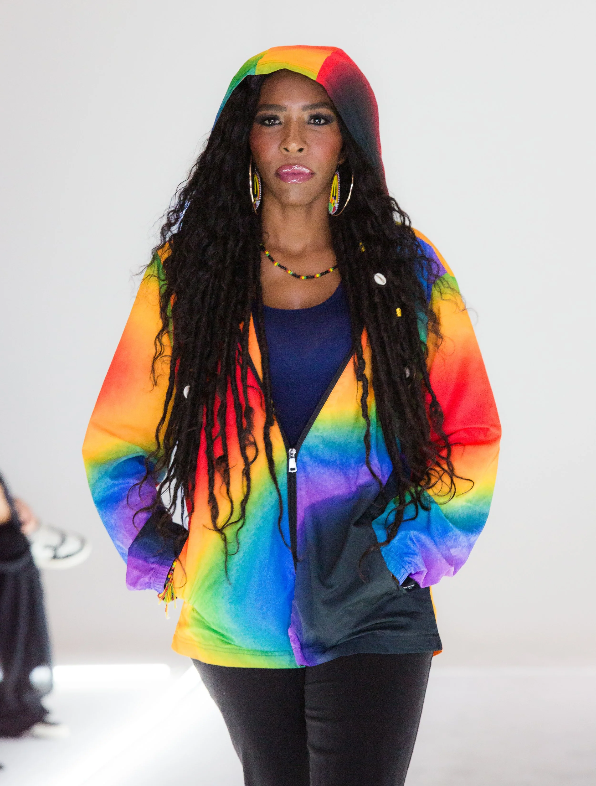 A woman with dark curly hair wearing a rainbow hoodie with matching earrings, necklace, and black pants.
