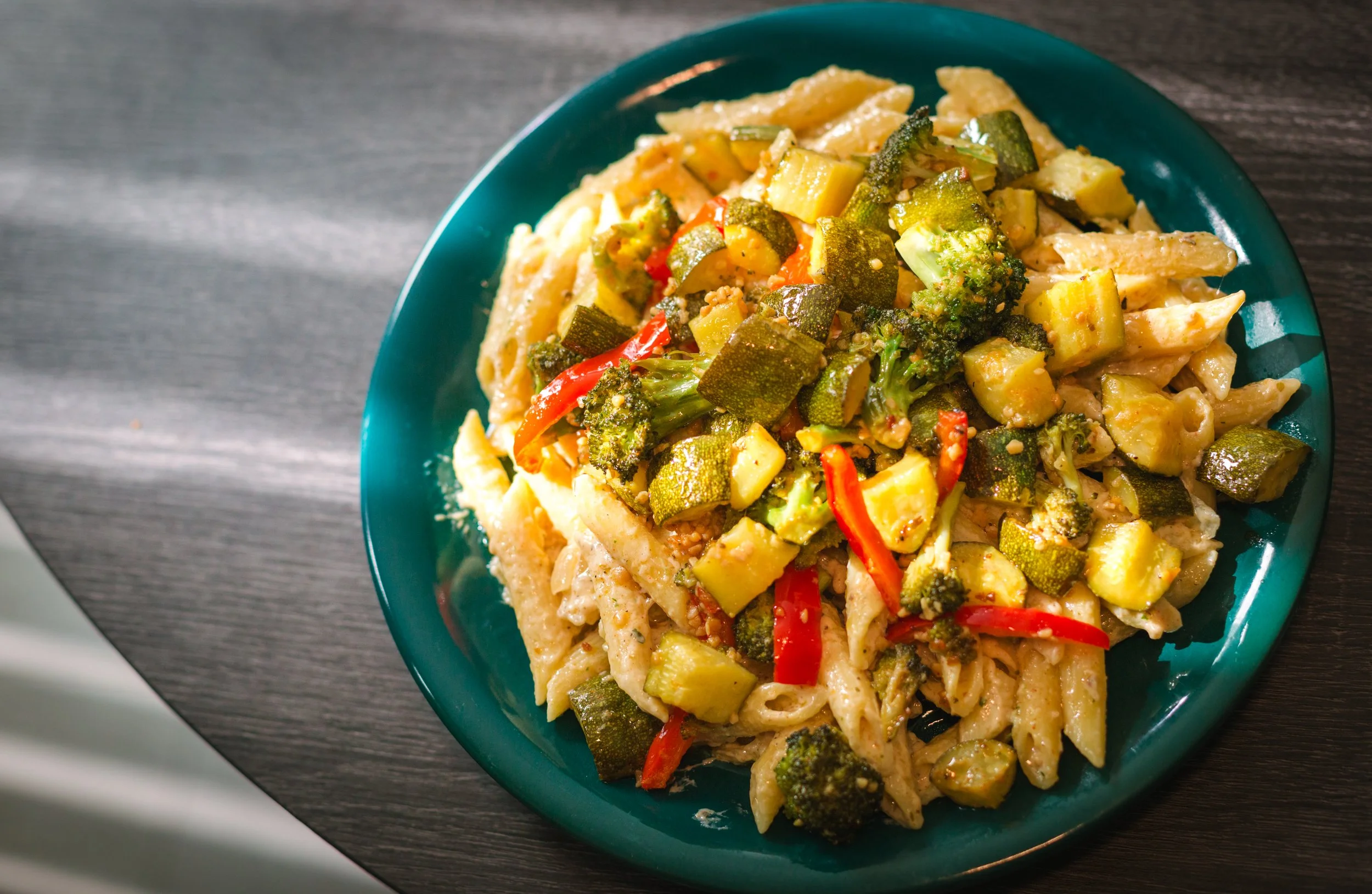 A plate of cooked pasta with mixed vegetables including broccoli, zucchini, and red bell peppers on a dark wooden surface.